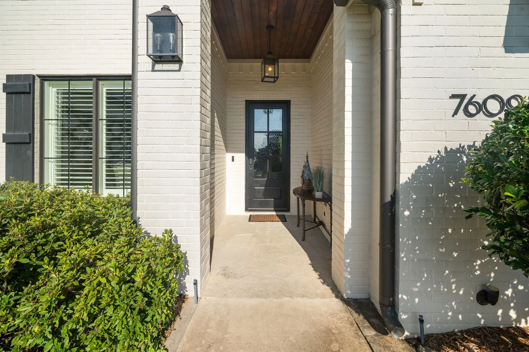 Covered entryway featuring a painted brick facade, wood-plank ceiling, black metal-framed front door with glass inlay, and structured exterior lantern lighting