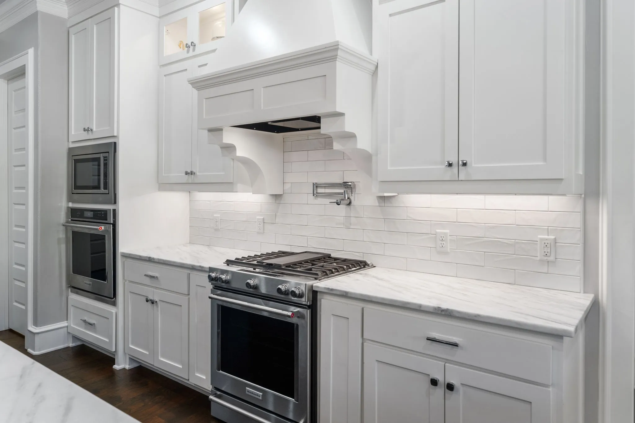 Kitchen featuring white shaker-style cabinetry, natural stone countertops, subway tile backsplash, stainless steel gas range with a custom decorative vent hood, and built-in wall oven and microwave