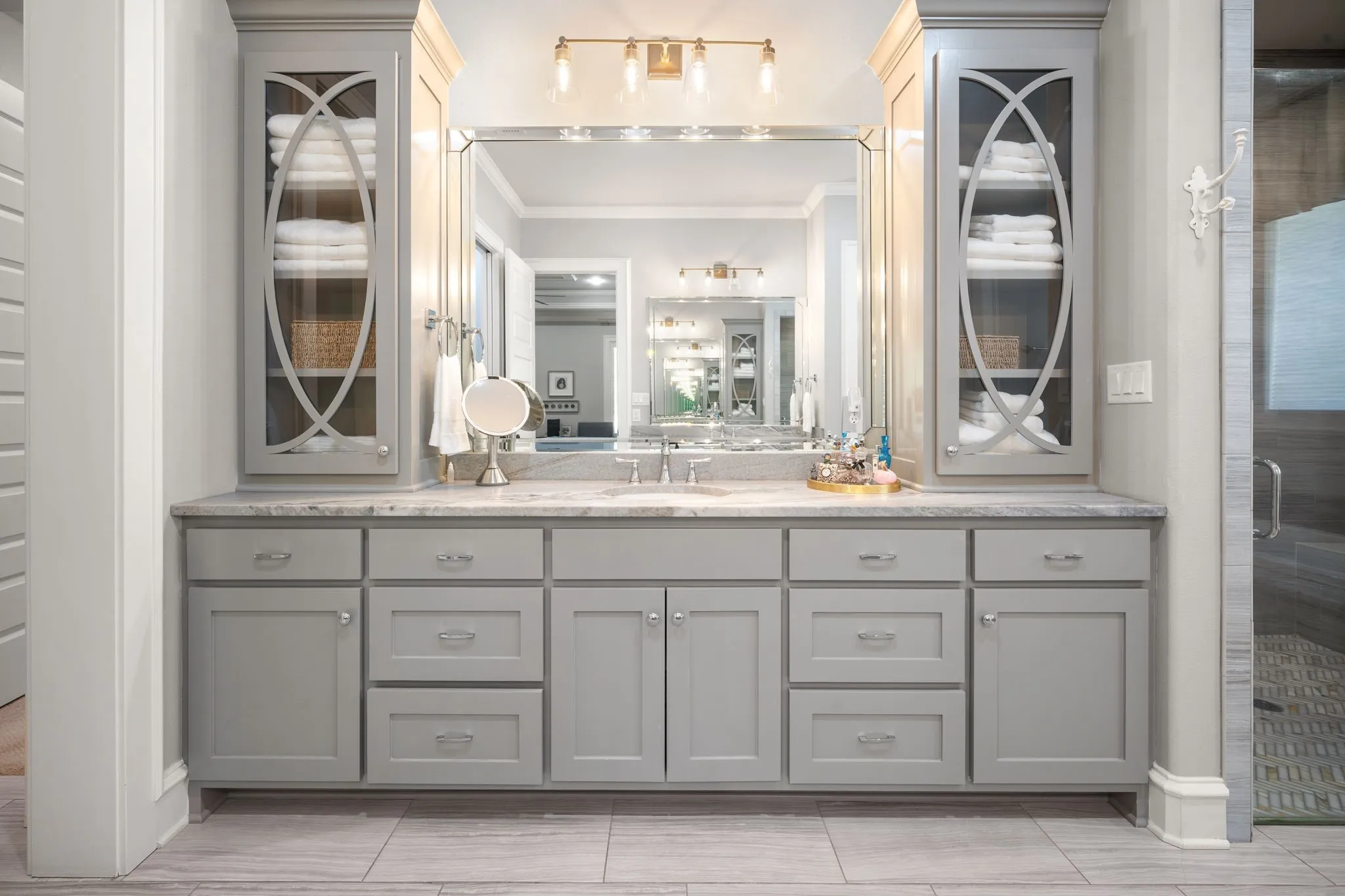 Dual-sink vanity featuring gray shaker-style cabinetry, integrated glass-front shelving, a natural stone countertop, and an oversized polished-edge mirror with multi-bulb vanity lighting