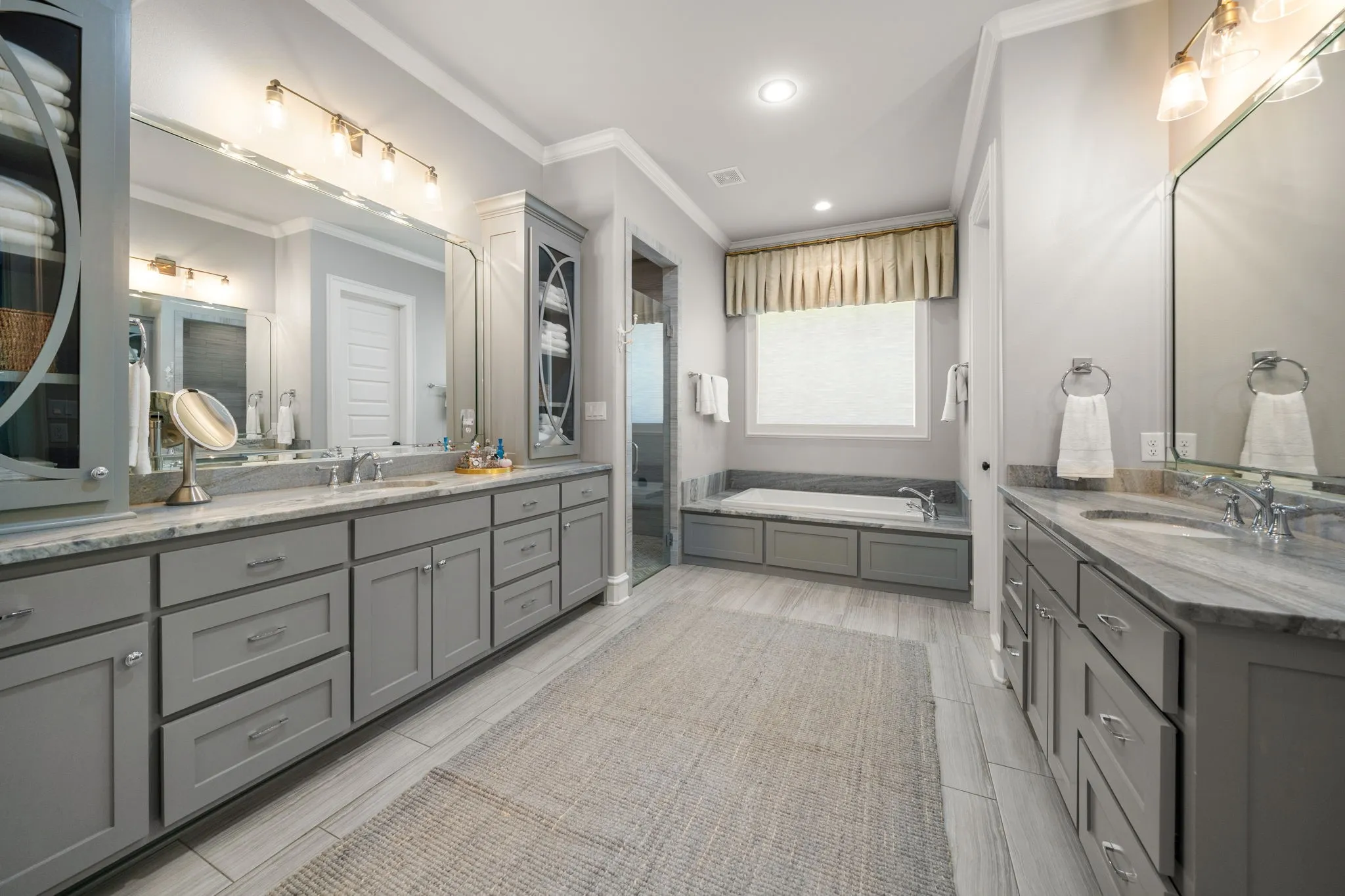 Dual gray cabinetry vanities with stone countertops, undermount sinks, full-width mirrors, deep soaking tub, and tile flooring