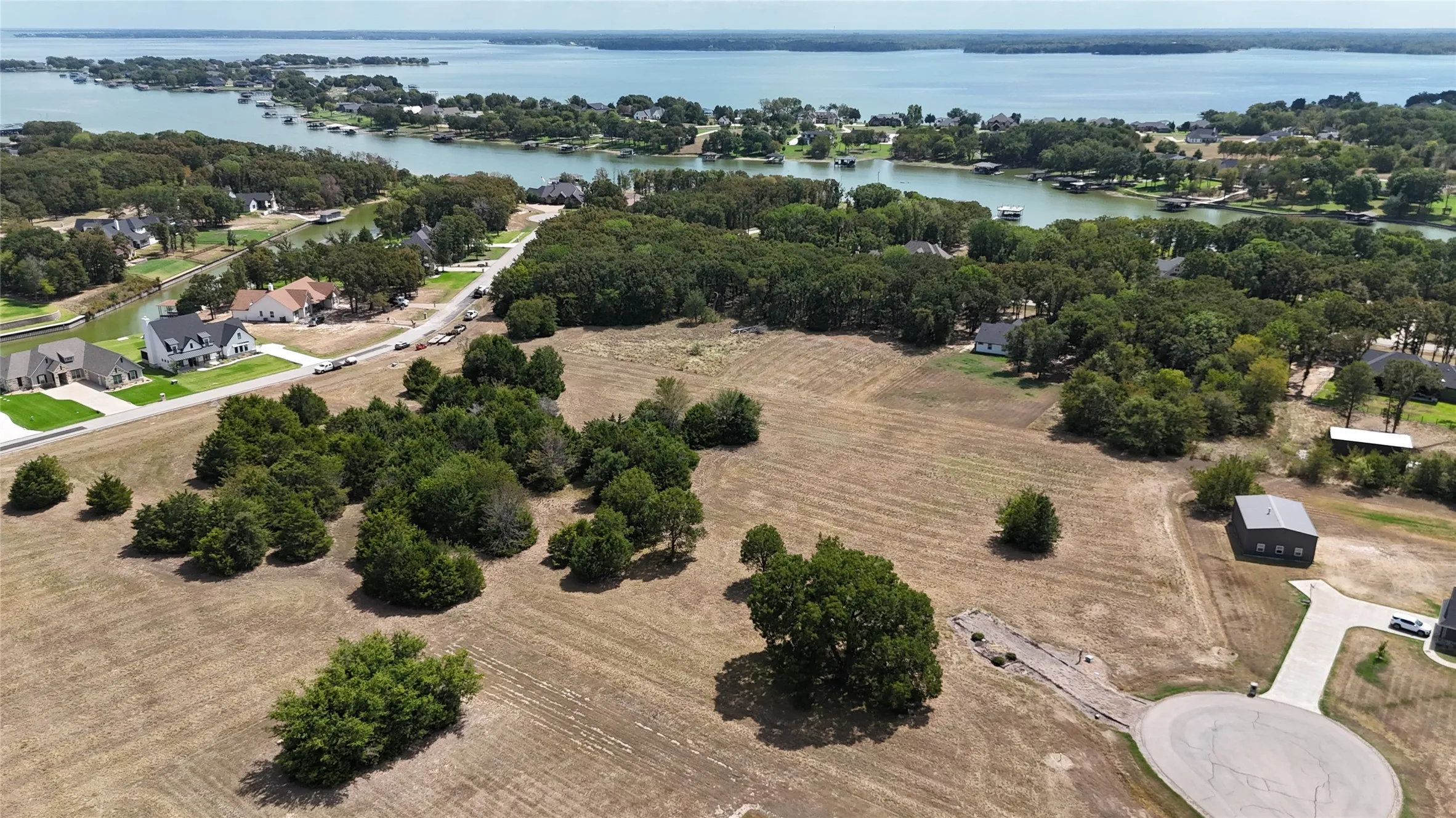 Expansive waterfront land parcel featuring mature trees, a paved cul-de-sac entrance, and a detached metal-roofed structure