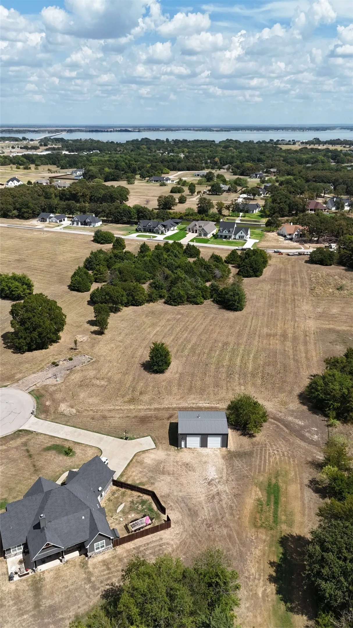 Aerial perspective featuring an expansive lot with mature tree clusters, a detached metal-roof garage, a paved driveway, and water frontage
