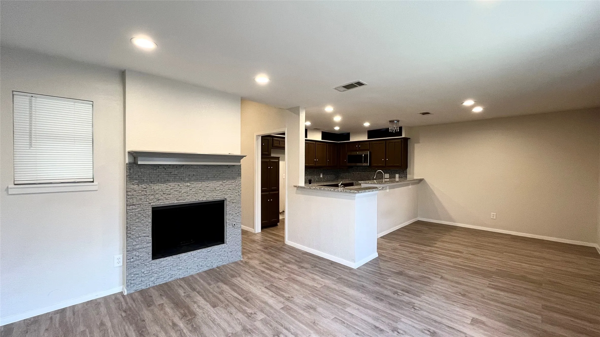 Fireplace with stacked-stone surround and mantel, wood-finish flooring, recessed lighting, and kitchen featuring dark cabinetry and a peninsula