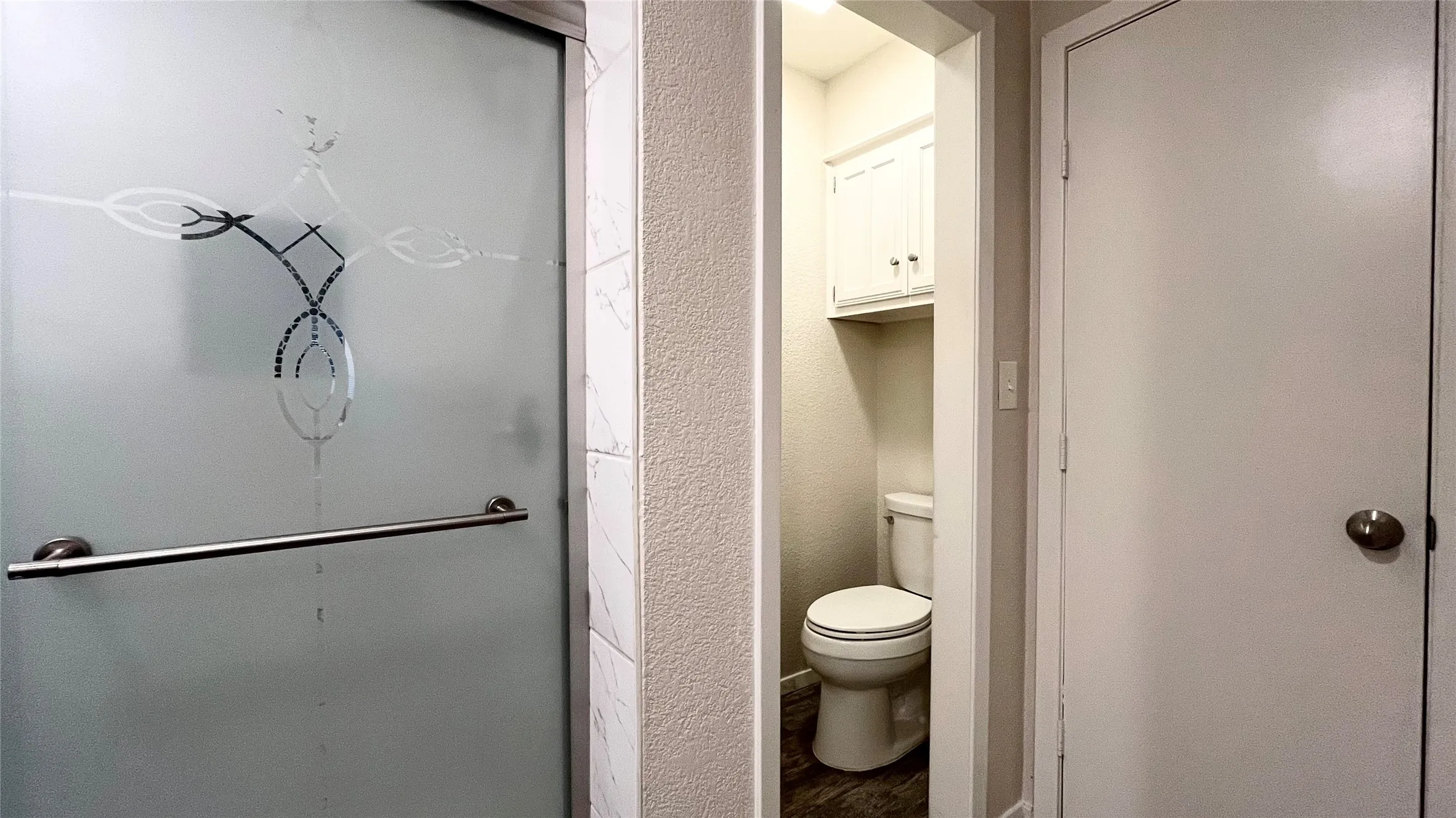 Frosted glass shower door with metallic handle, neutral wall tile, two-piece toilet, and built-in upper wall cabinetry