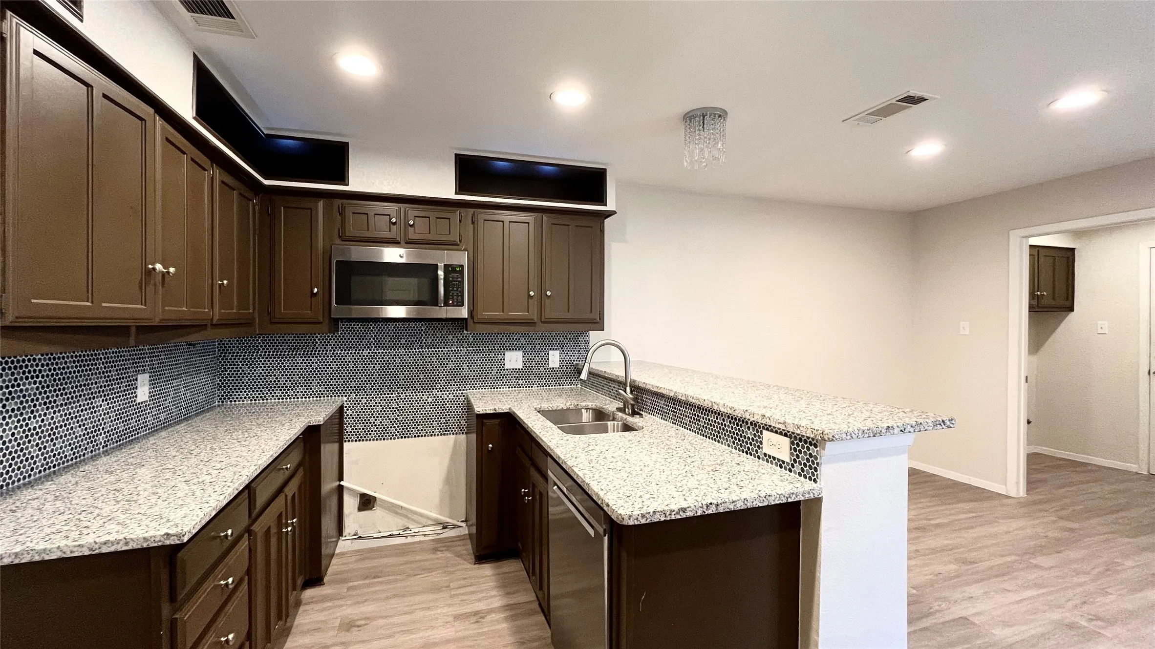 Kitchen featuring dark wood cabinetry, speckled stone-finish countertops, geometric tile backsplash, stainless steel microwave, and wood-finish flooring