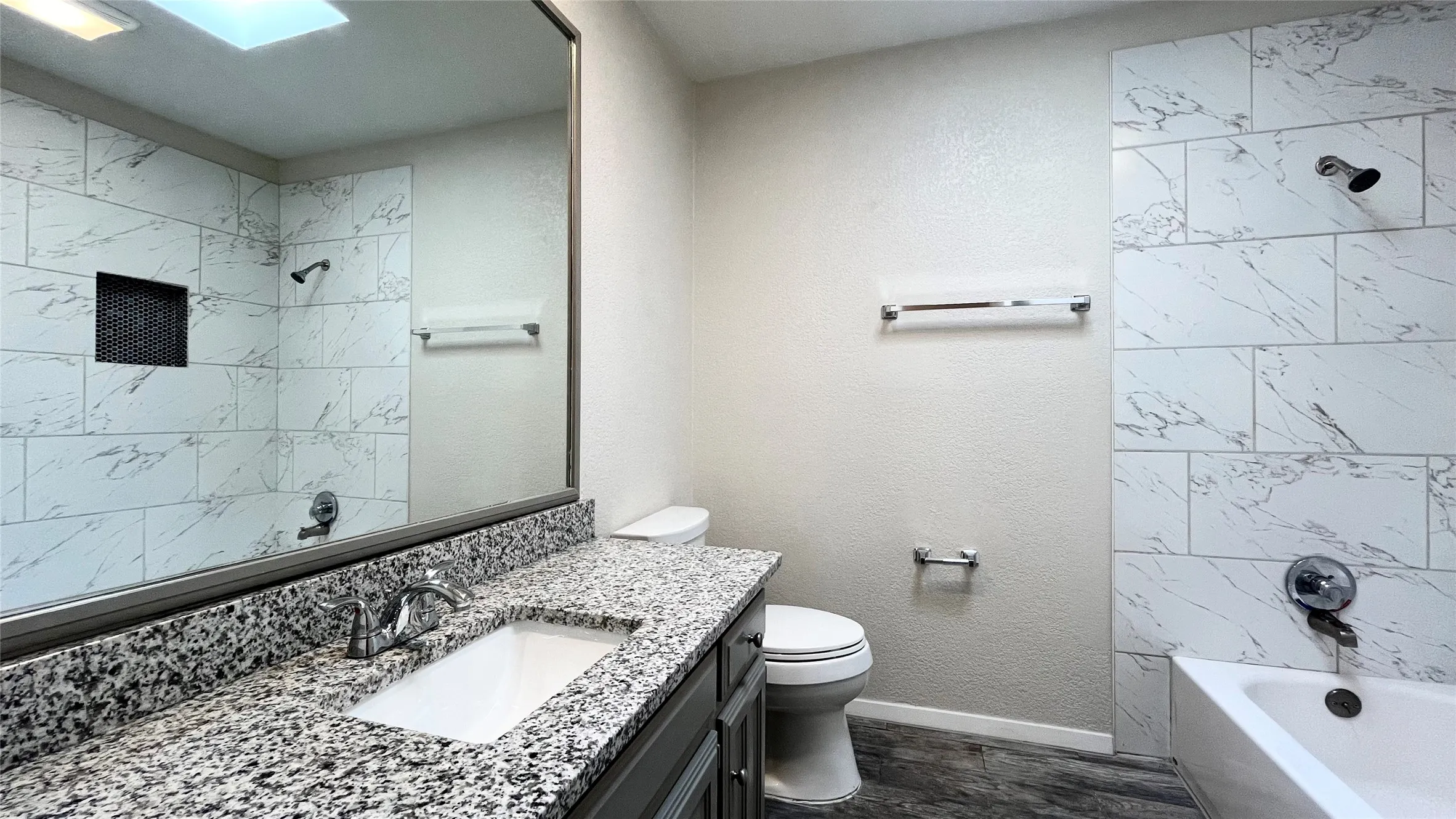Full-width vanity with a stone-finish countertop, undermount sink, large wall-mounted mirror, bathtub featuring large-format tile surround, and built-in shower niche