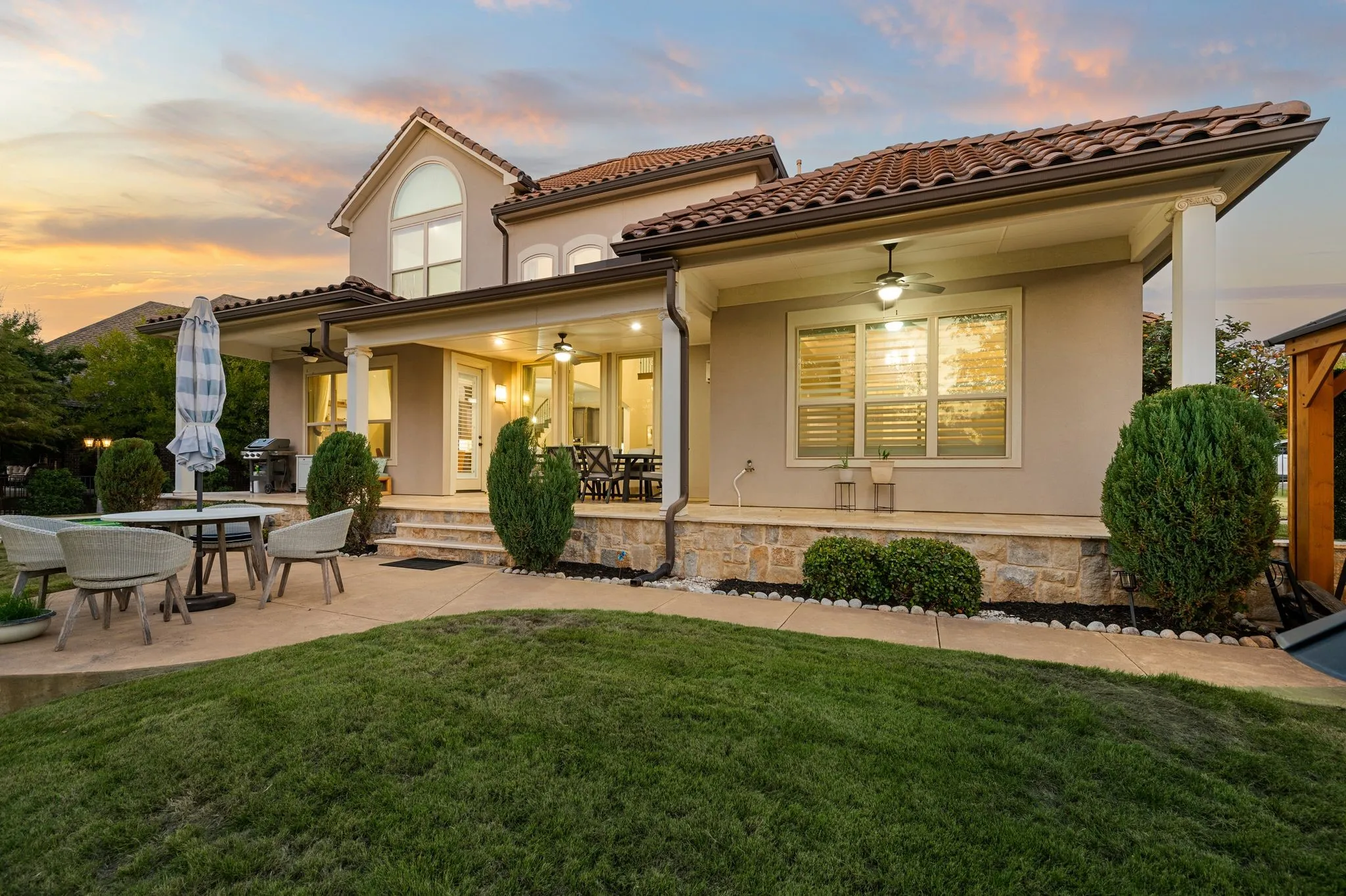 Covered patio with a stone-base facade, tile roof, and multiple ceiling fans, featuring a concrete walkway and manicured lawn