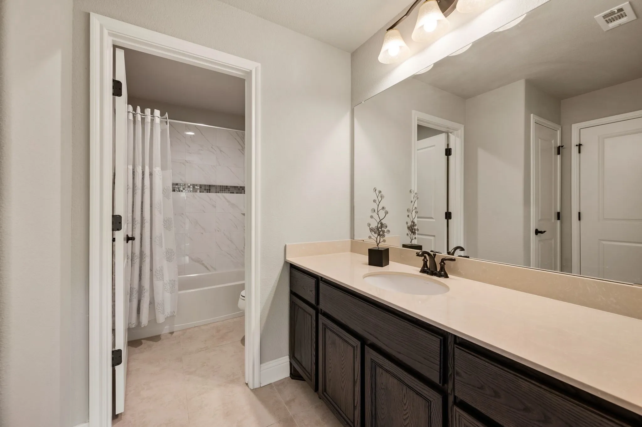 Bathroom featuring a dark wood-finish vanity, light stone-surface countertop, an oval undermount sink with dual-handle faucet, full-width wall mirror, and a tub-shower combination with tile-finish surround