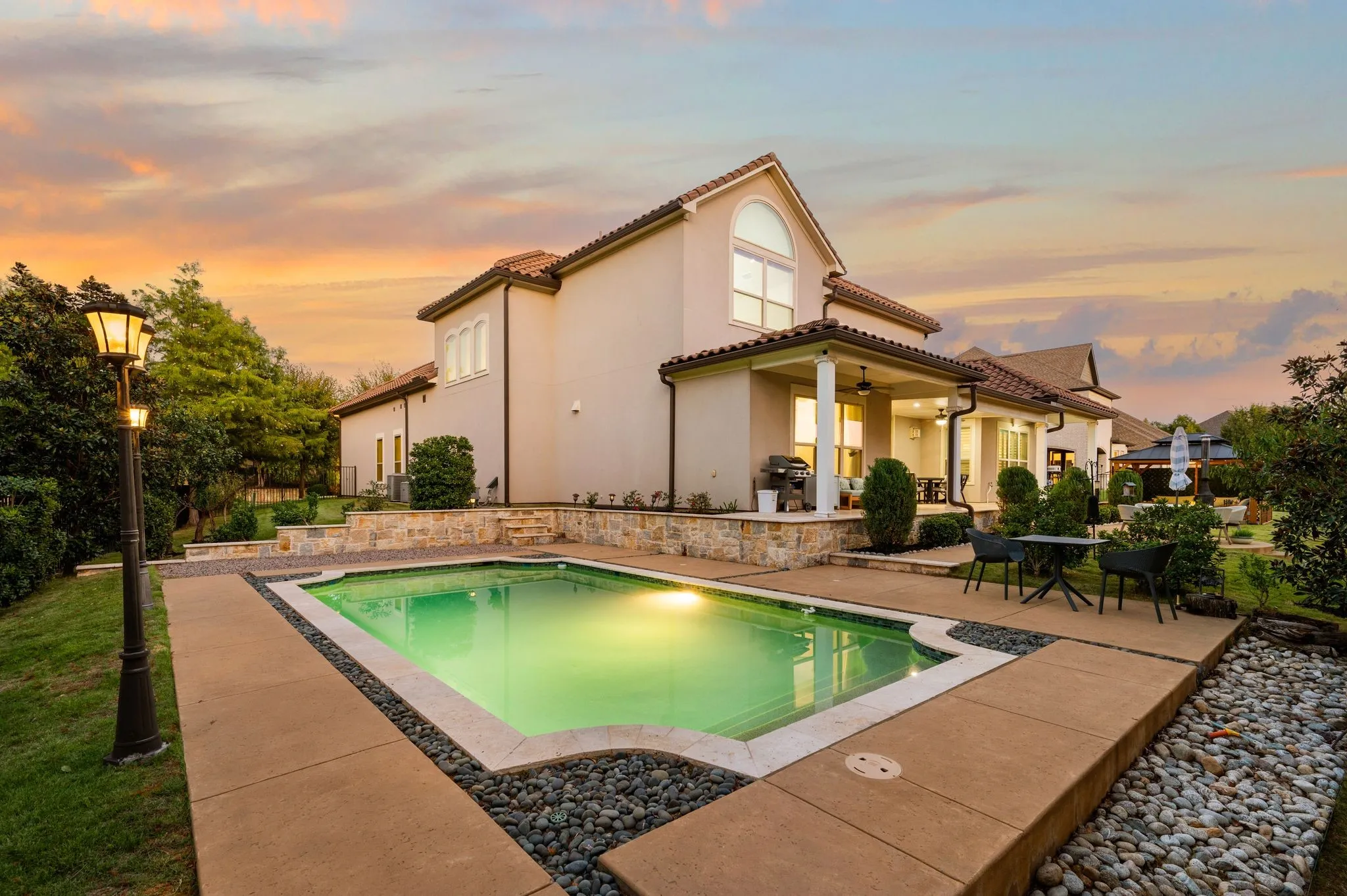 Exterior rear elevation featuring a stucco facade, covered patio, in-ground swimming pool with stone coping, and decorative rock hardscaping