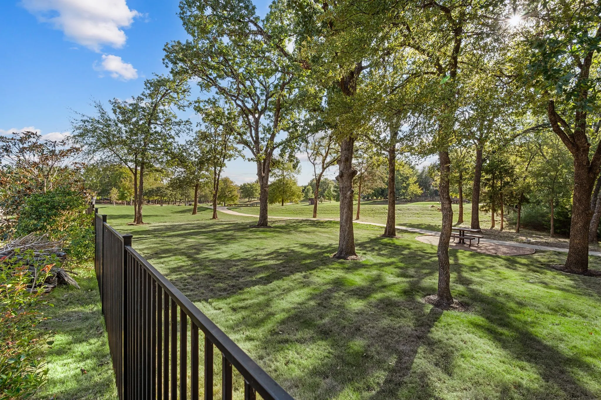 Community walking trails featuring mature trees, a manicured lawn, a black metal perimeter fence, and a stone-paver patio area