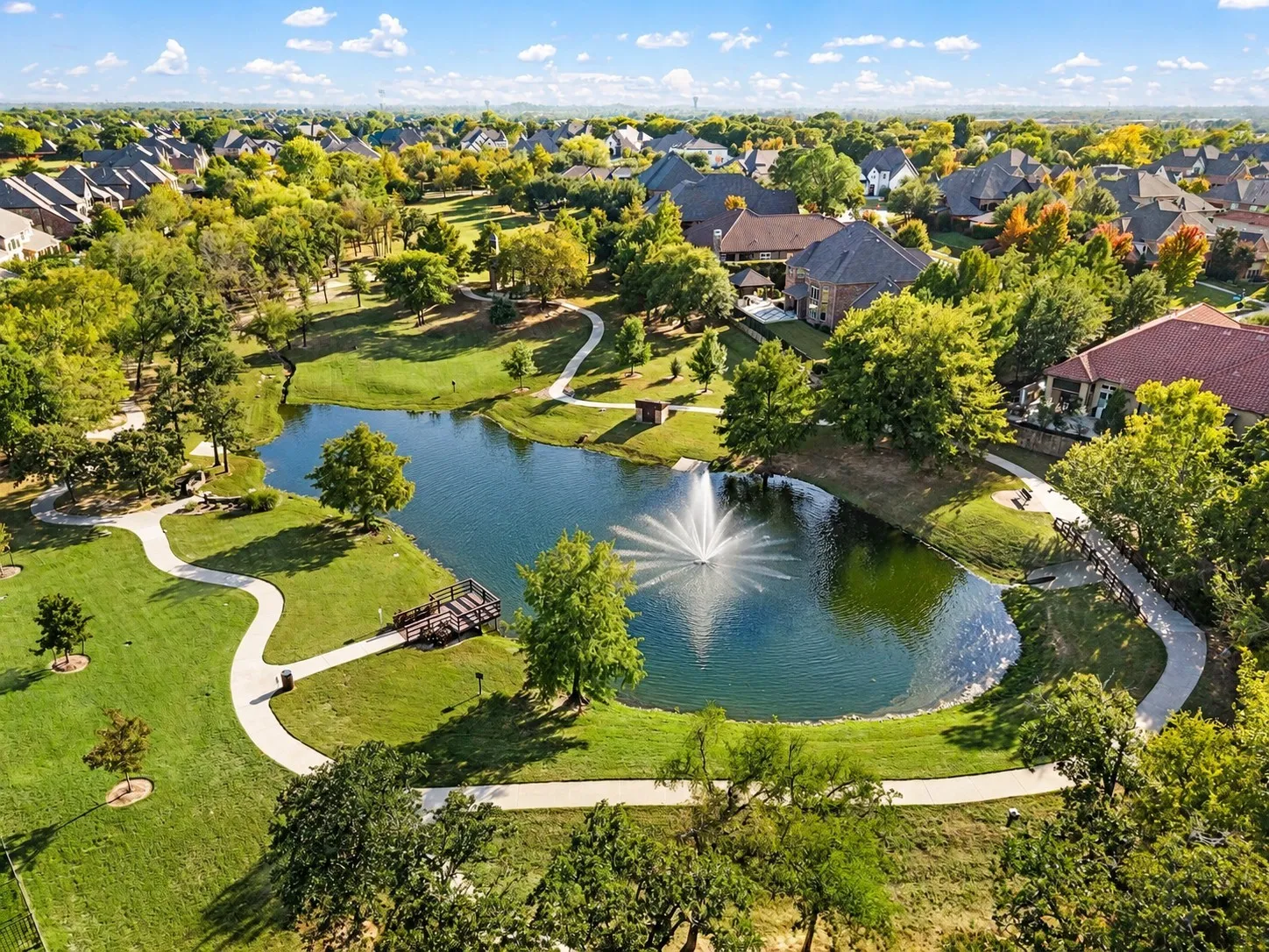 Community grounds featuring a central pond, a multi-tiered water fountain, paved walking paths, a wooden pedestrian bridge, and an elevated observation deck