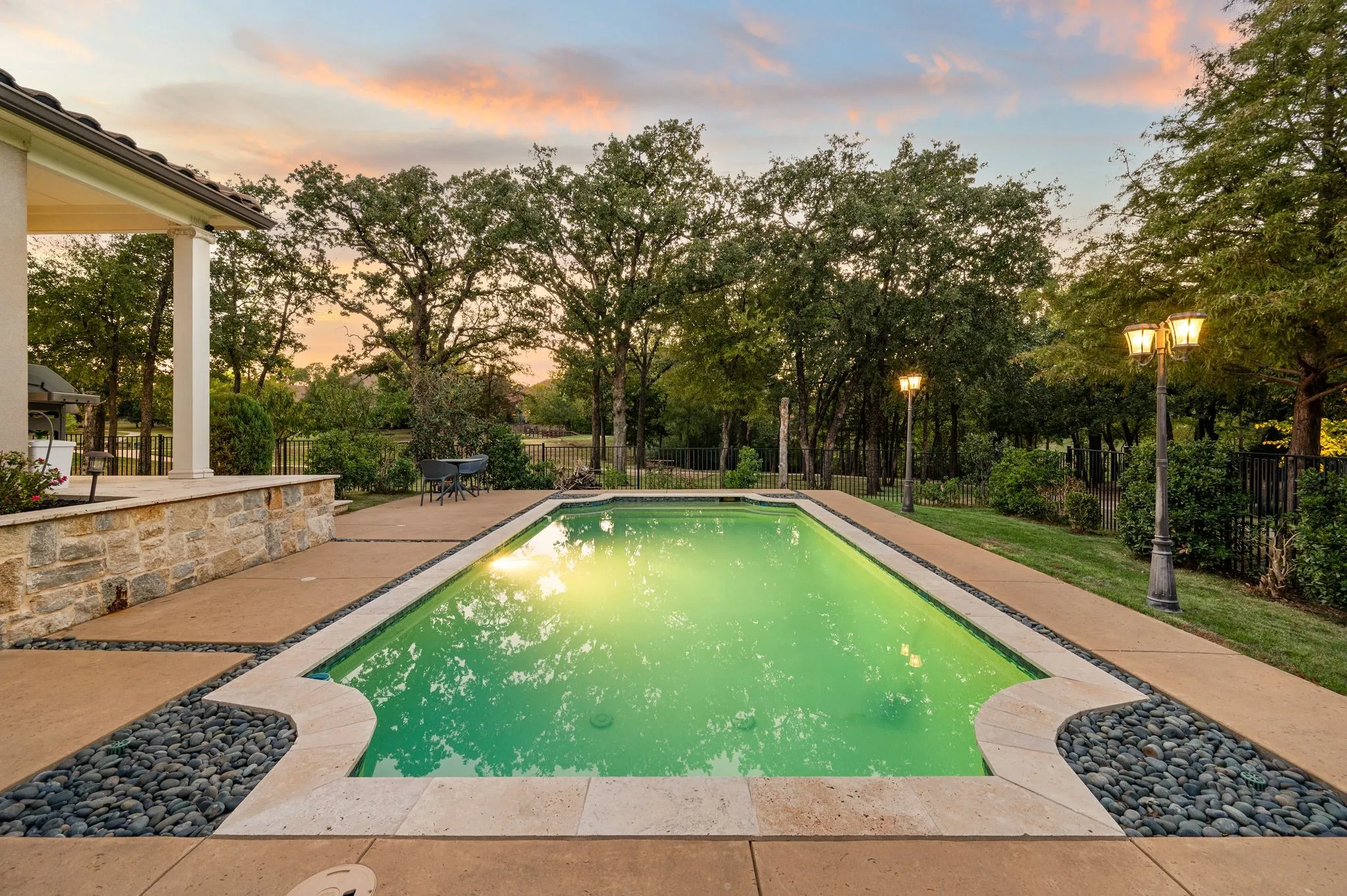 Inground swimming pool with stone coping, concrete patio, stone retaining wall, mature trees, and decorative light posts