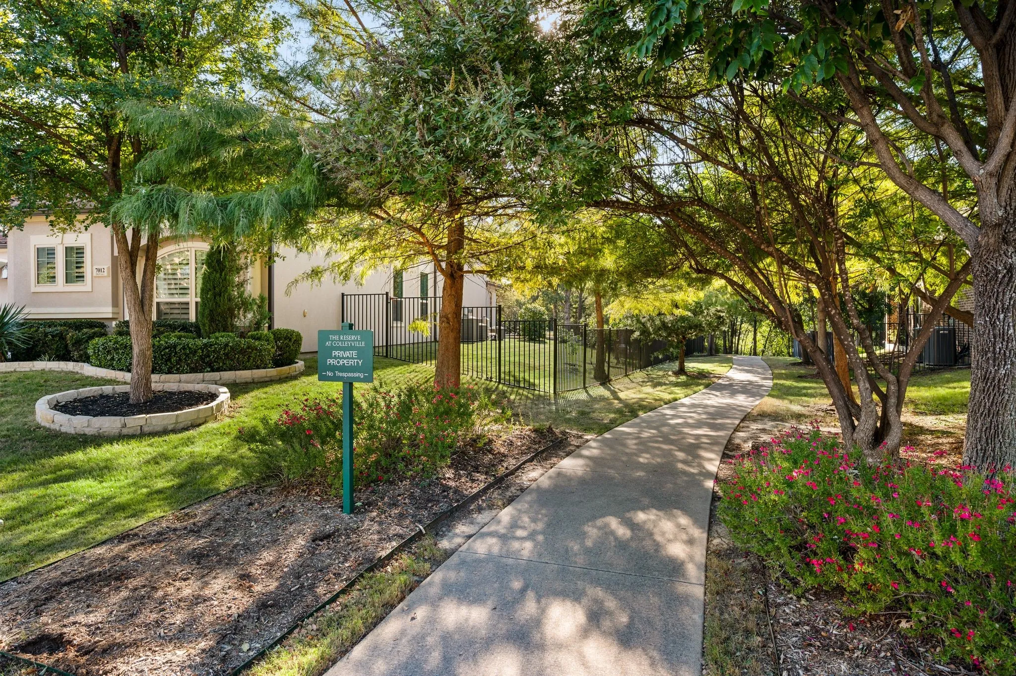 Concrete walkway bordered by mature landscaping, stone-edged garden beds, and metal perimeter fencing