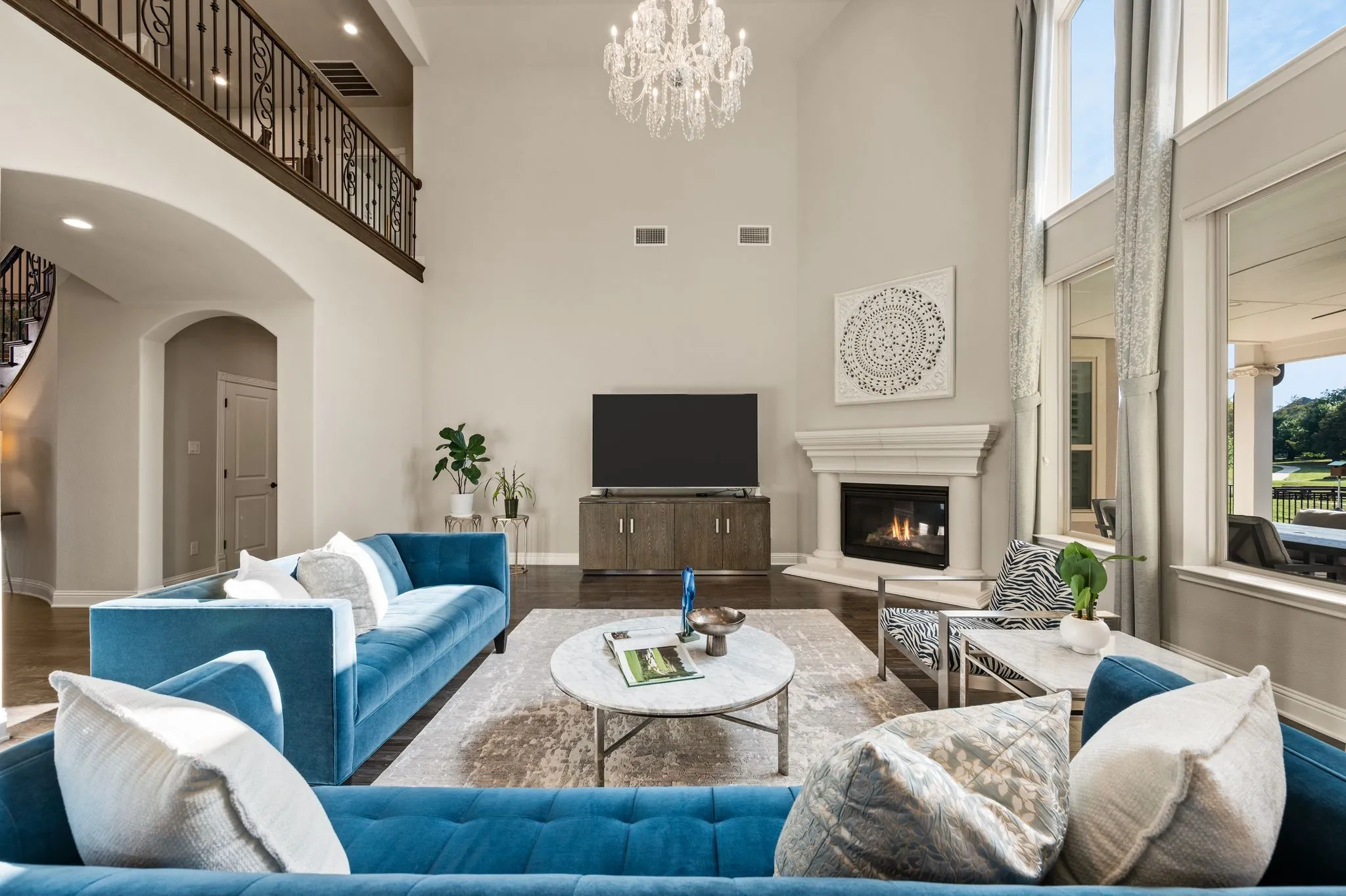Double-height ceiling, floor-to-ceiling windows, corner gas fireplace with decorative mantel, crystal chandelier, and wrought iron balcony railing