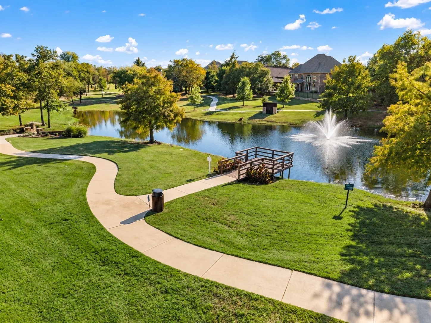 Community grounds featuring a pond with a central fountain, a wooden observation dock, and concrete walking paths set amidst landscaped green space with mature trees
