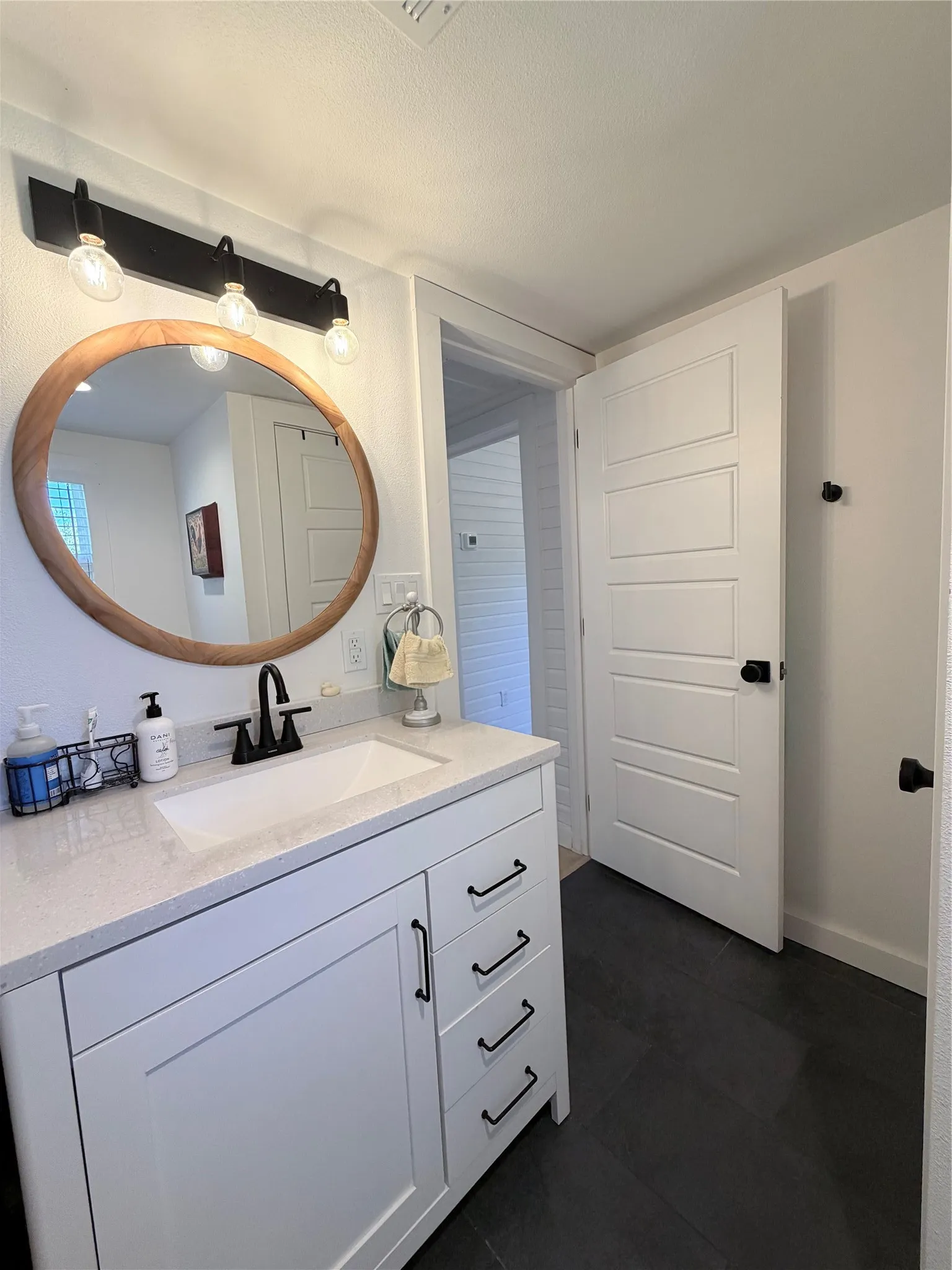 White cabinetry with matte black hardware, stone-finish countertop, integrated rectangular sink with black faucet, round wood-framed mirror, and three-light matte black vanity fixture