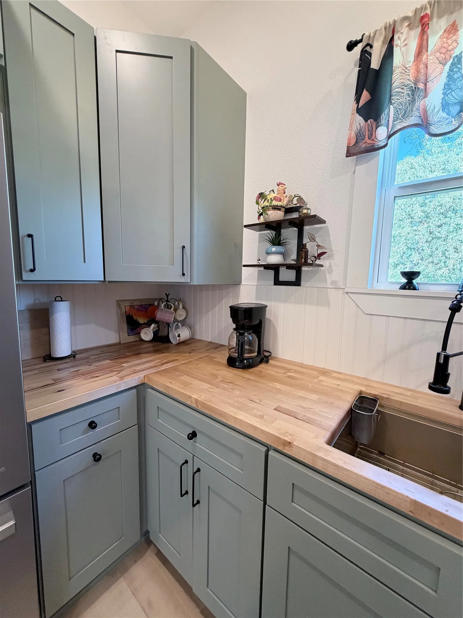 Sage green cabinetry featuring Shaker-style doors, butcher-block countertops, a stainless steel drop-in sink with matte black hardware, and beadboard wall paneling