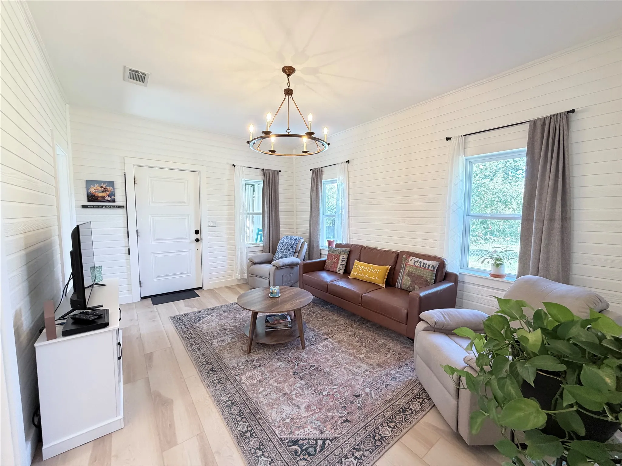 Interior space featuring horizontal wood-plank wall paneling, light-tone wood-finish flooring, a wrought-iron candelabra chandelier, and multiple windows with white trim