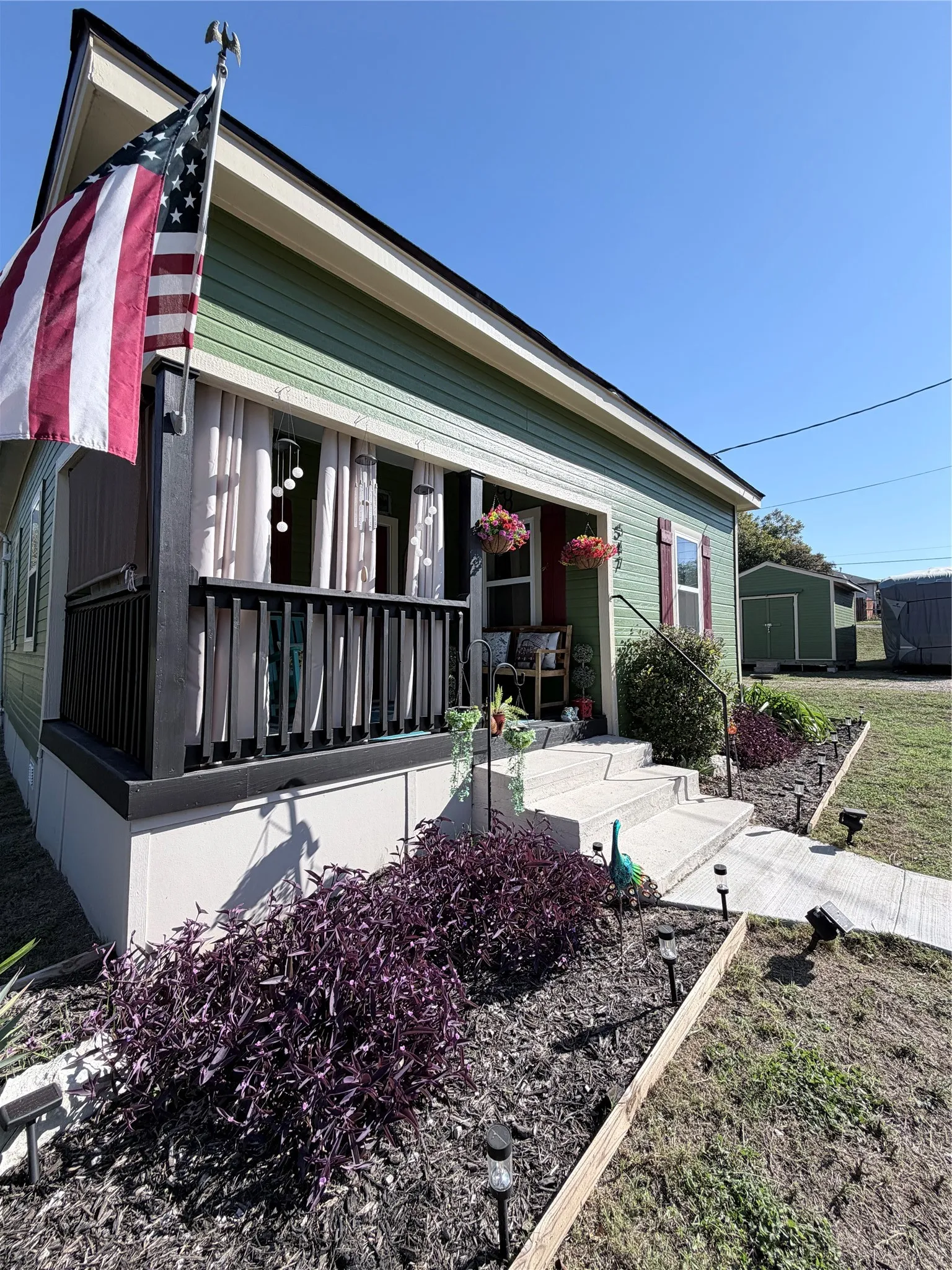 Exterior featuring horizontal siding, a covered front porch with a black railing, concrete steps, landscaped garden beds with mulch, and an attached storage structure