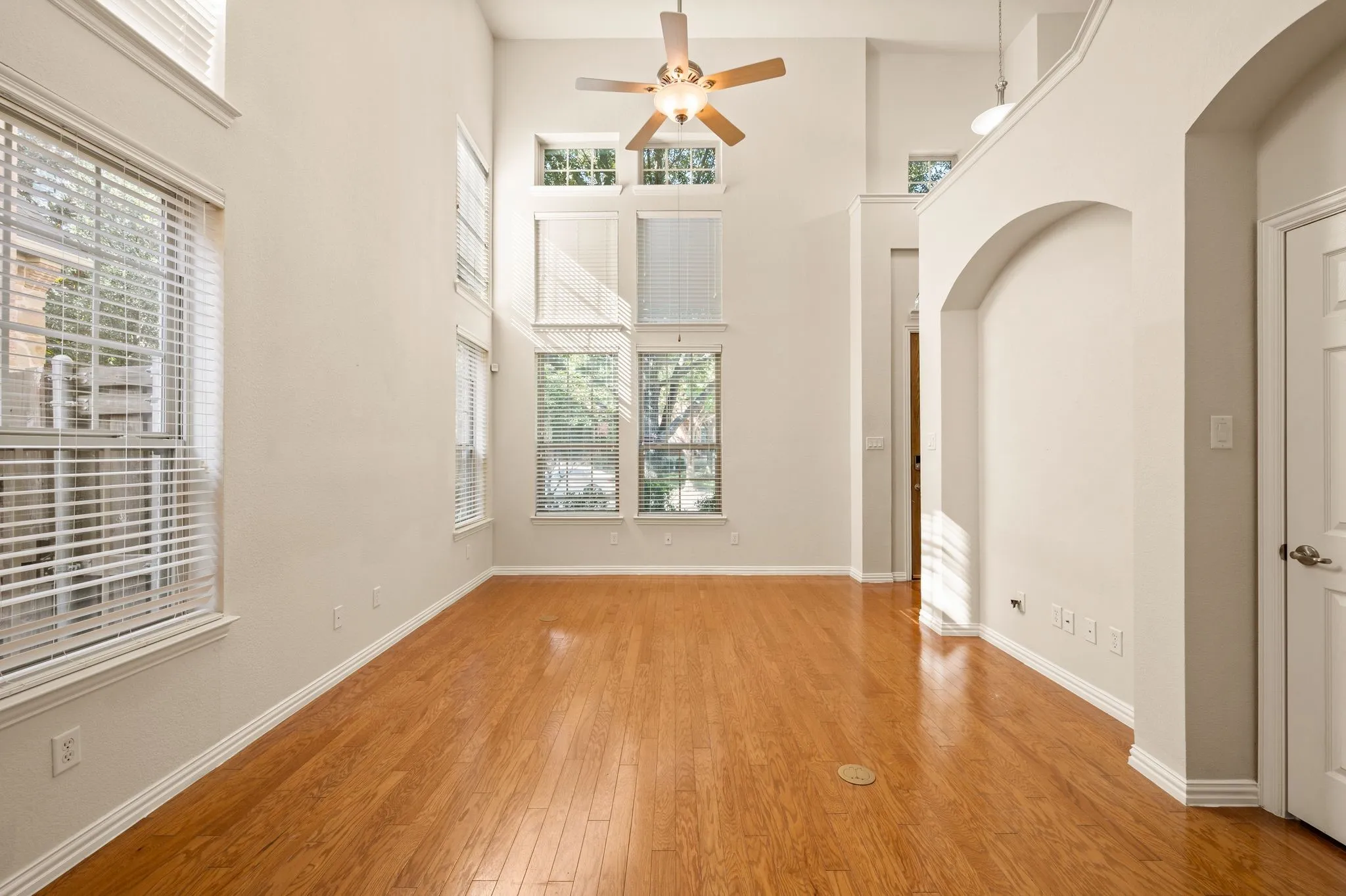 High-ceiling interior featuring wood-finish flooring, multi-level windows, architectural arched alcove, and a central ceiling fan with integrated lighting