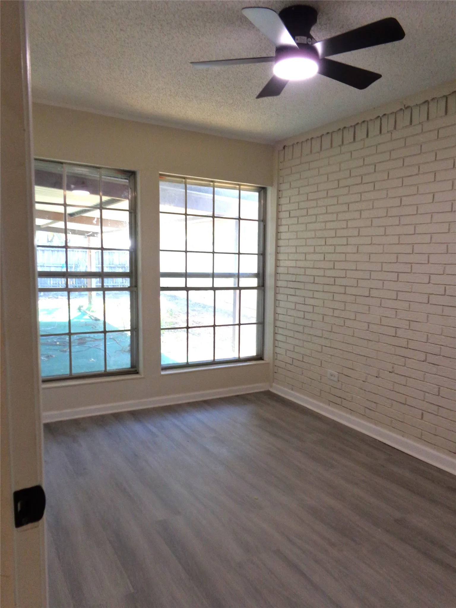 This could be a 4th Bedroom, Nursery, or Office. Interior space featuring wood-finish flooring, a painted brick accent wall, multi-pane windows, and a modern ceiling fan with integrated lighting