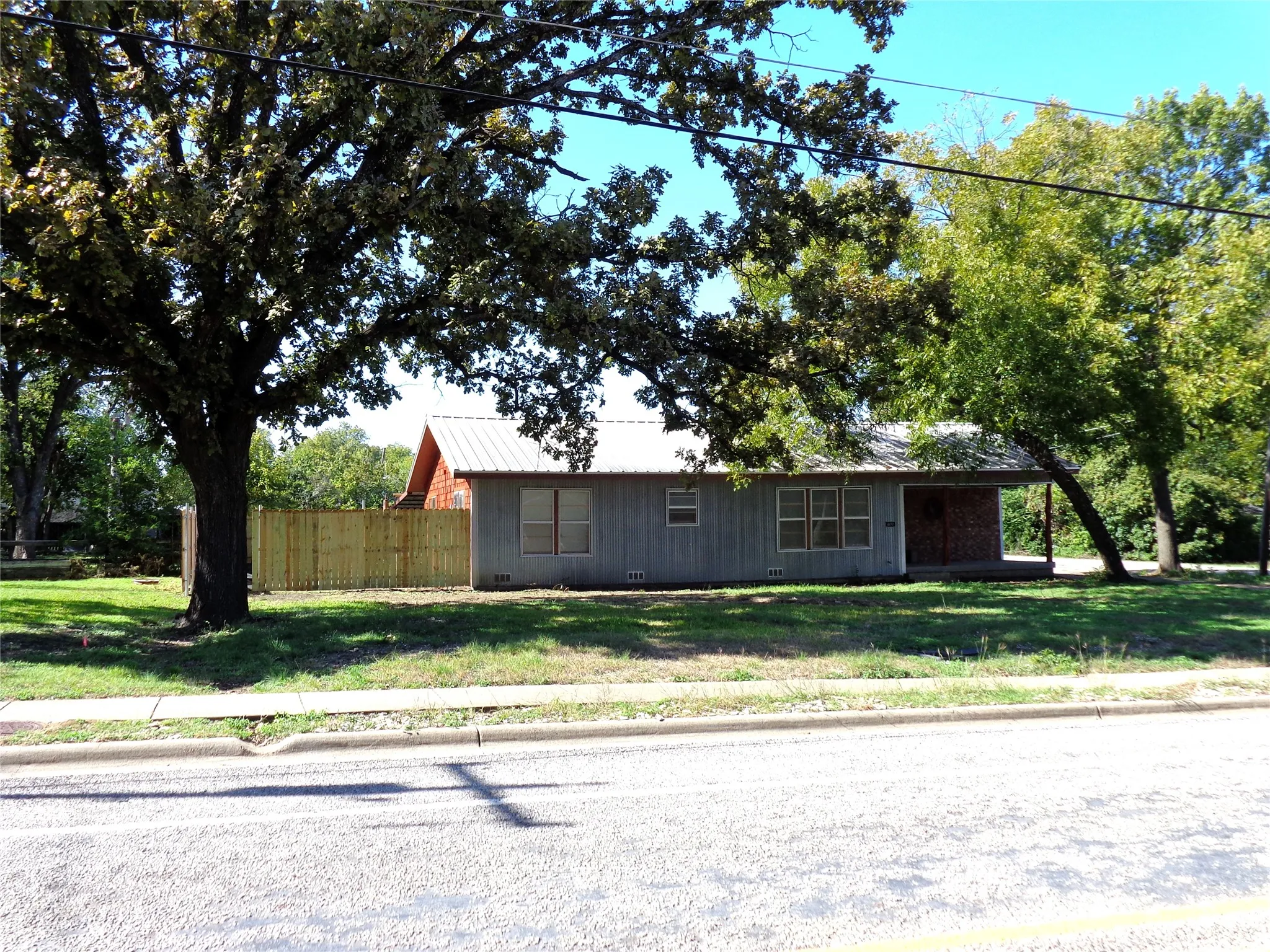 Single-story exterior featuring light-toned wood siding, a metal roof, a recessed porch with brick accents, and vertical wood privacy fencing