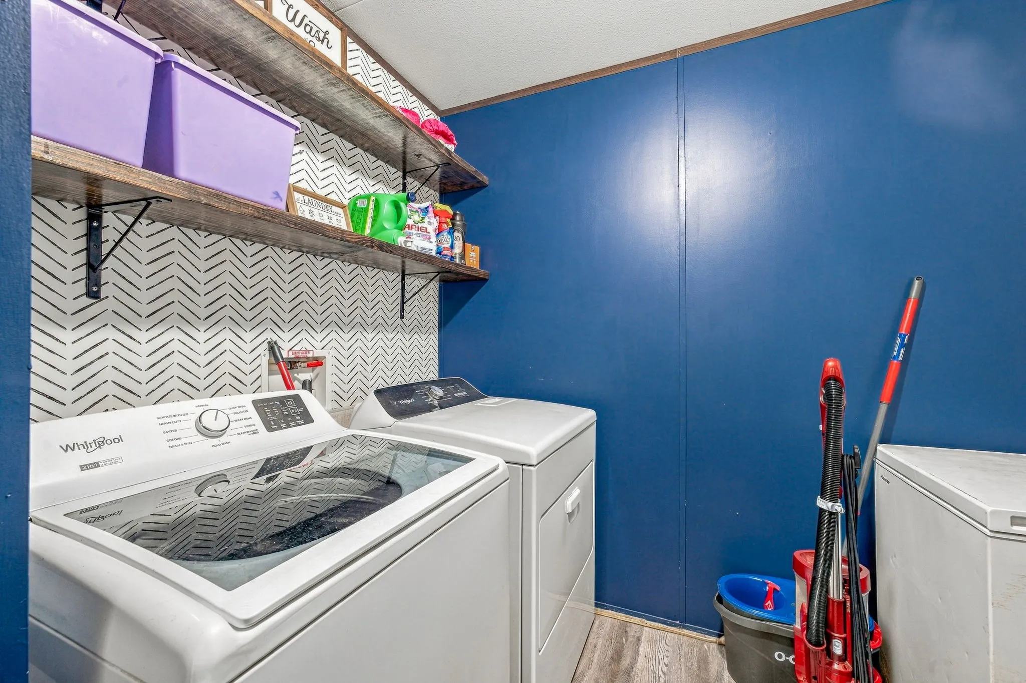Laundry area featuring wood-finish flooring, herringbone-patterned accent wall, and multi-tier open shelving with metal supports