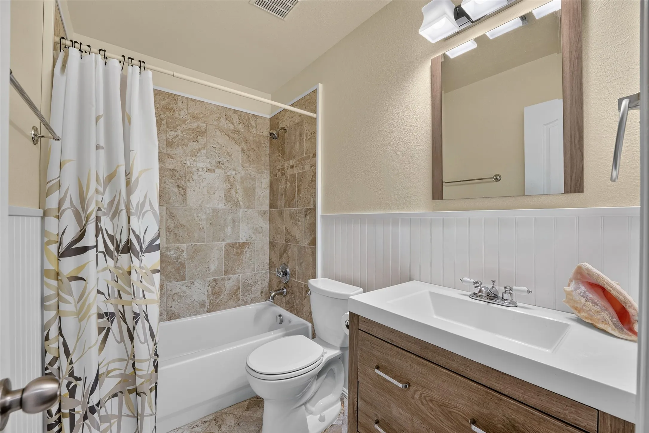 Super cute completely remodeled bathroom featuring a tub/shower combination with tile surround and flooring,  wood vanity with rectangular sink, framed mirror, white beadboard wainscoting and decorative mirror and coordinated fixtures.