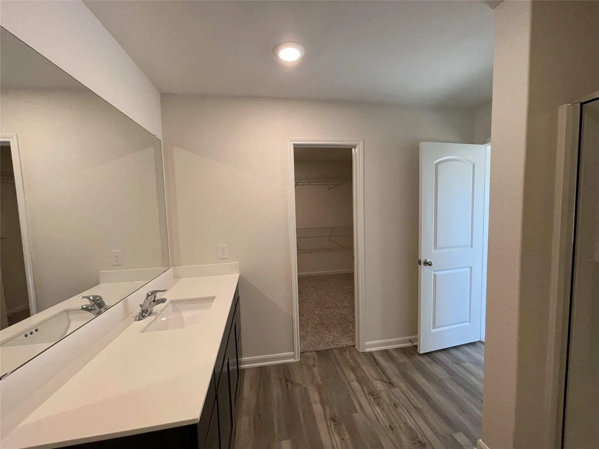 Bathroom featuring a dual-sink vanity with a solid-surface countertop, full-width wall mirror, wood-finish flooring, and a walk-in closet