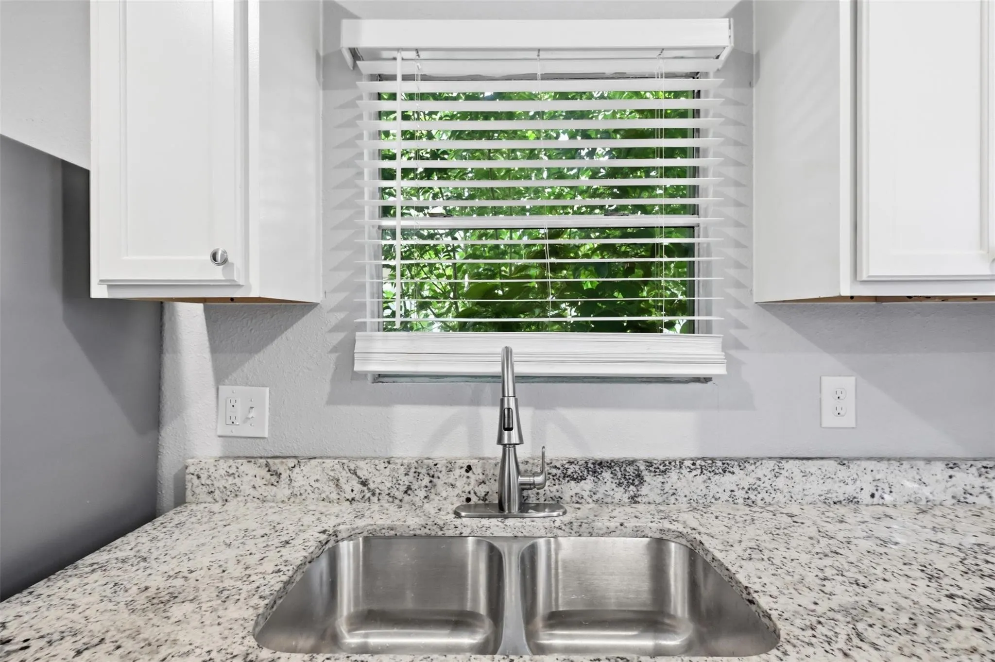 Double basin stainless steel sink with a high-arch pull-down faucet, light-toned stone-finish countertop, white cabinetry, and horizontal window blinds