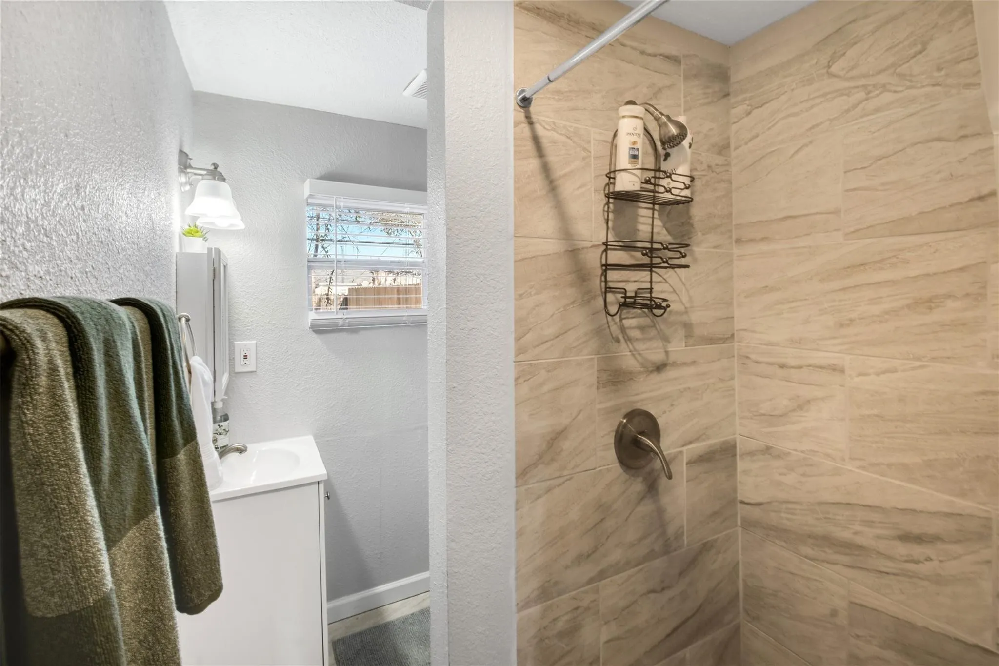 Bathroom featuring a shower stall with neutral-toned, large-format tile surround, single-handle brushed metal faucet, wall-mounted light fixture, and a white cabinet vanity with a built-in sink