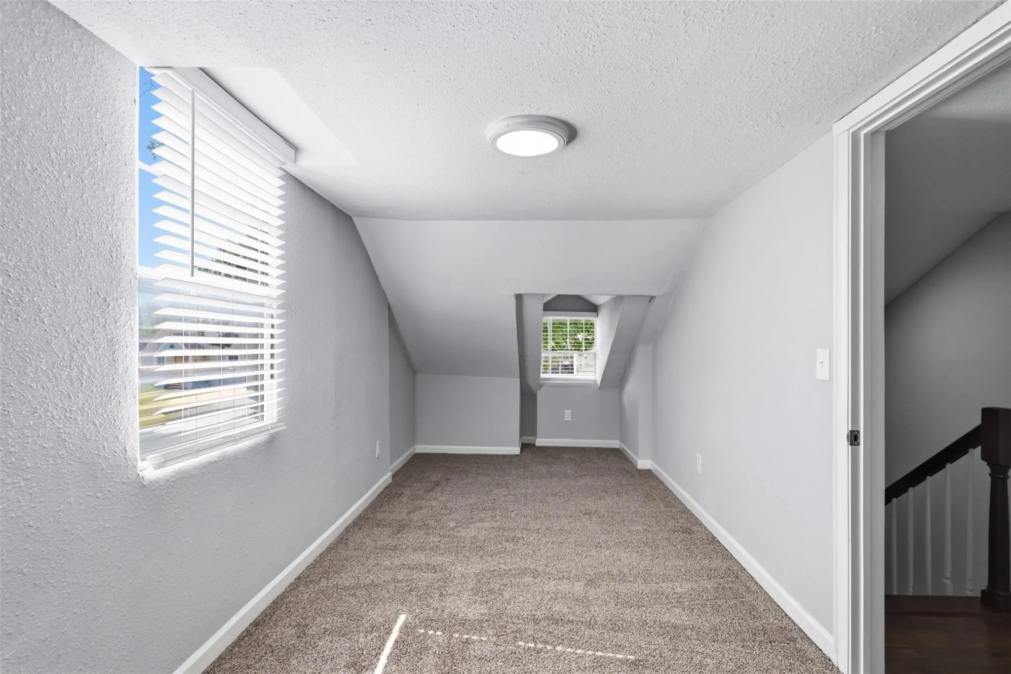 Neutral-toned interior room featuring slanted ceiling lines, wall-to-wall carpeting, and multiple windows with horizontal blinds