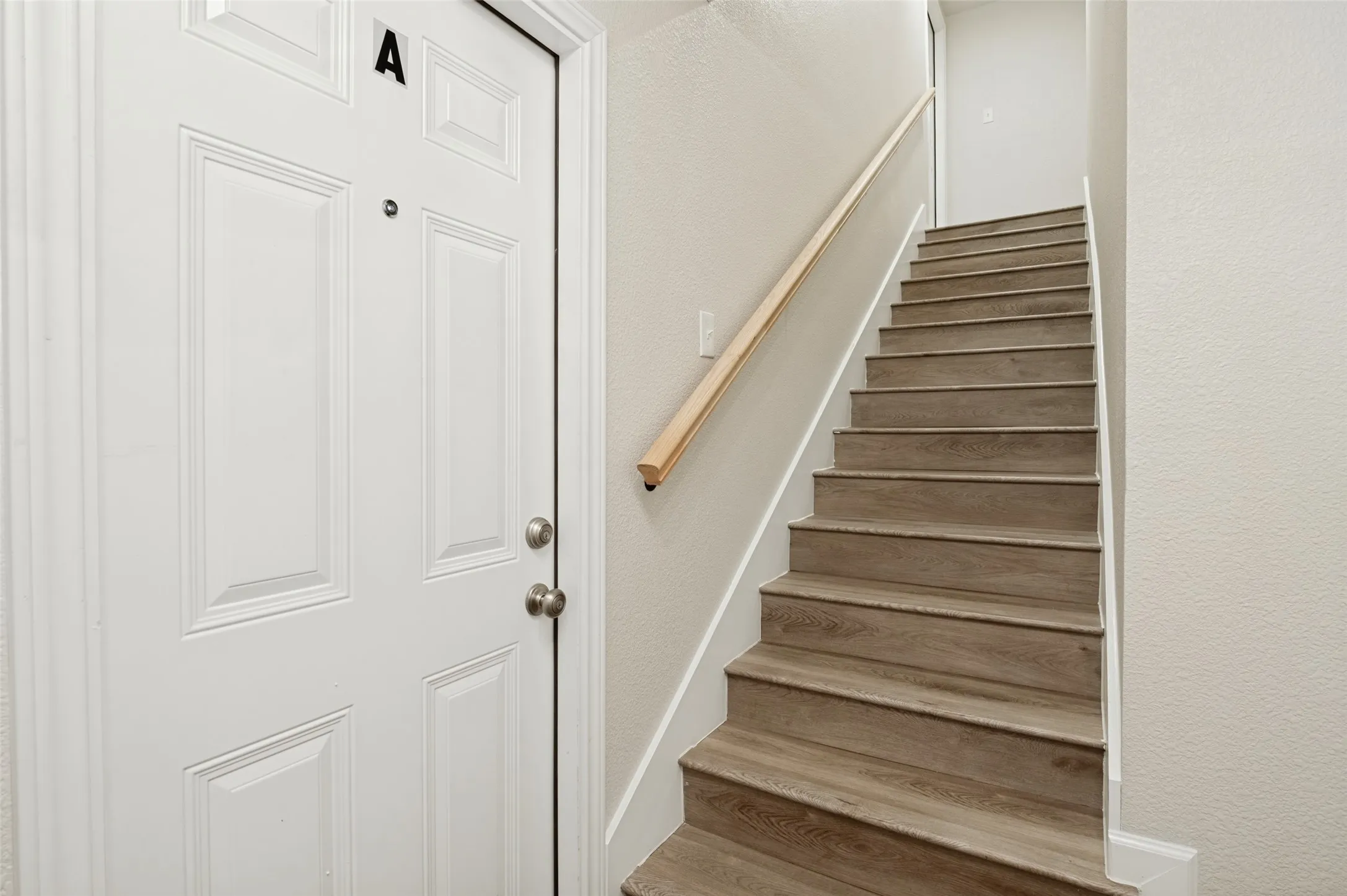 White paneled door with integrated deadbolt and peephole, accompanied by a staircase featuring wood-finish treads and a light-toned wood handrail