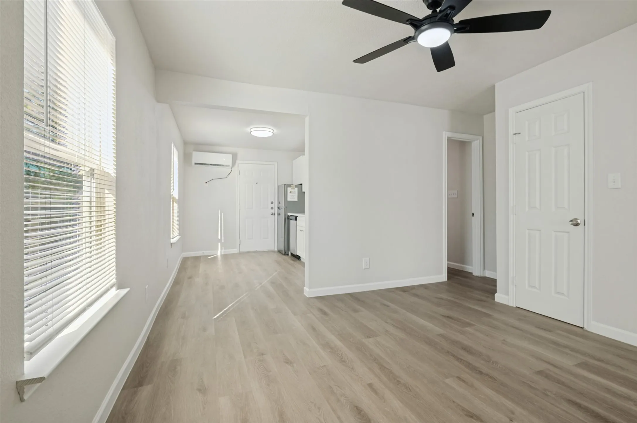 Wood-finish flooring, matte black ceiling fan with integrated lighting, wall-mounted ductless air conditioning unit, and white six-panel interior doors