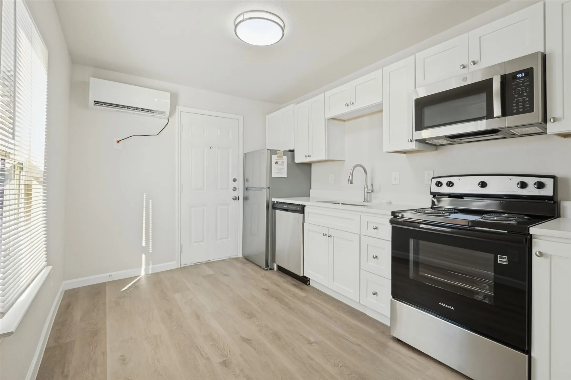 Kitchen featuring white shaker-style cabinetry, wood-finish flooring, stainless steel appliances, and a wall-mounted ductless heating and cooling unit