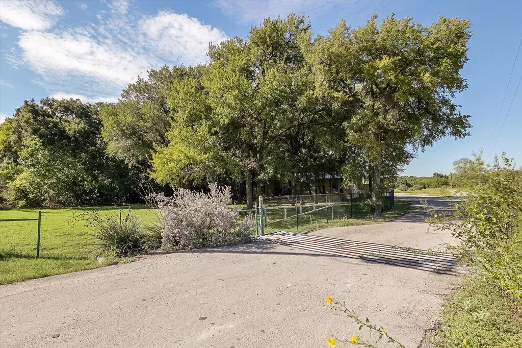 Paved road frontage featuring a gated entrance, open grassy acreage, and mature tree canopy