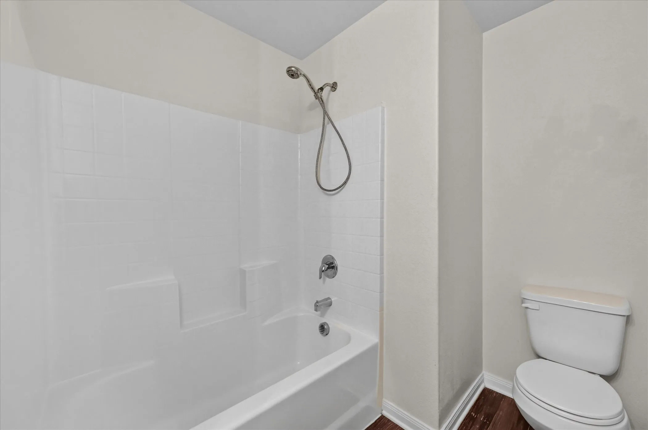 White acrylic tub and shower combination with a chrome handheld shower head, matching single-handle faucet, and wood-finish flooring