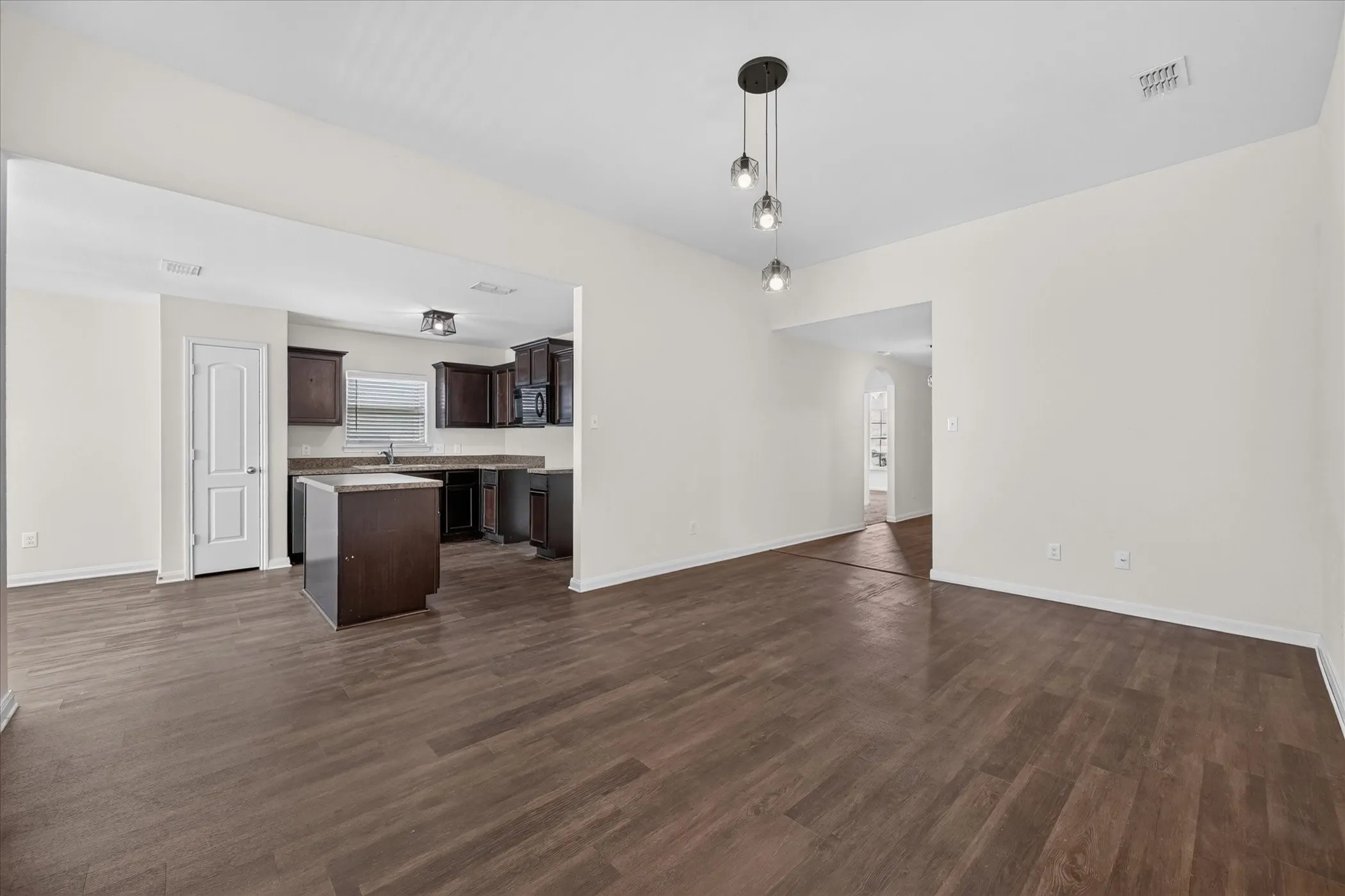 Open-concept living space with wood-finish flooring, three-globe pendant lighting, dark wood-finish cabinetry, center kitchen island, and neutral wall tones