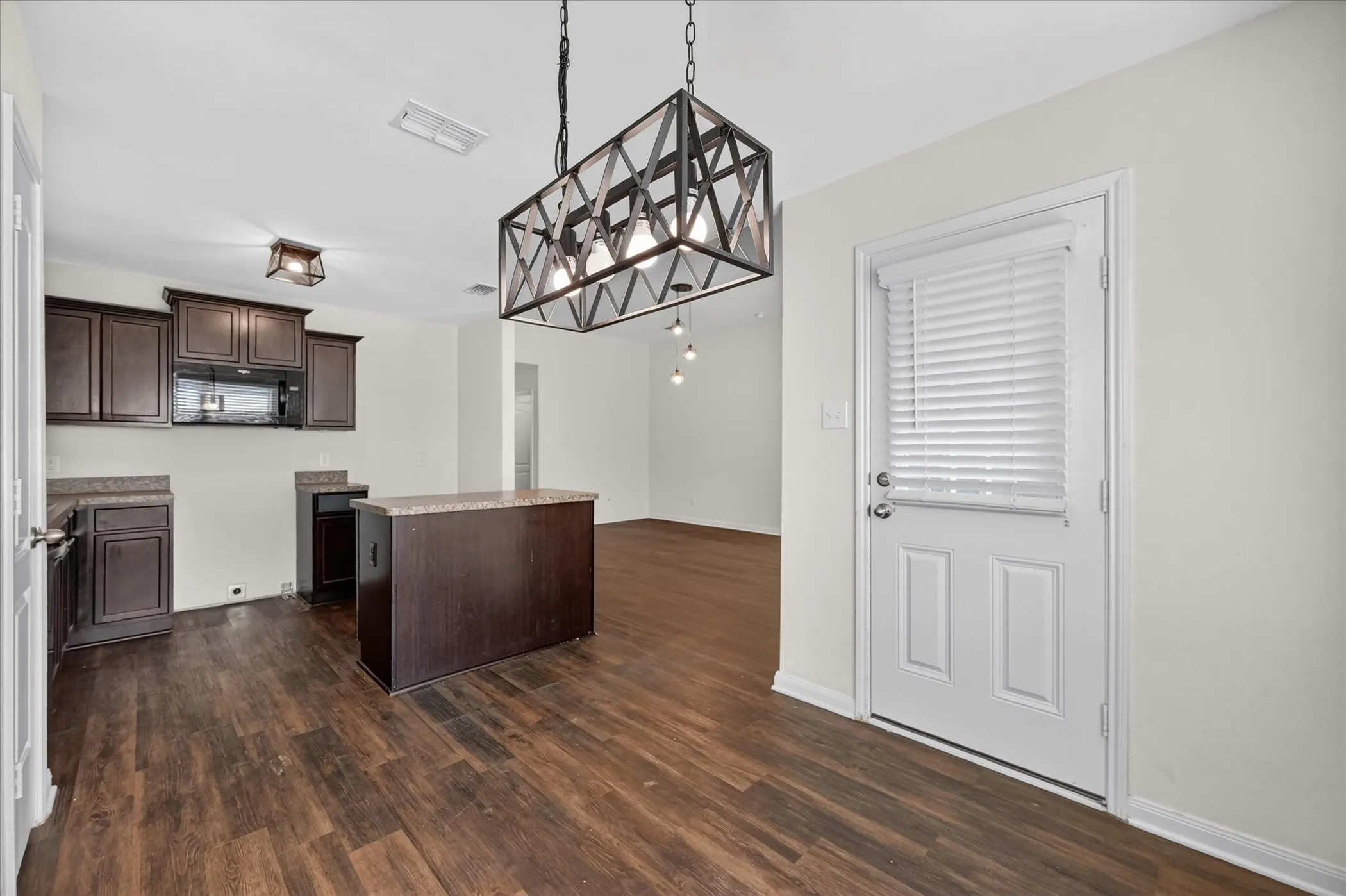 Dark-finish cabinetry, wood-finish flooring, central kitchen island, geometric metal-frame pendant lighting, and an exterior door with integrated blinds
