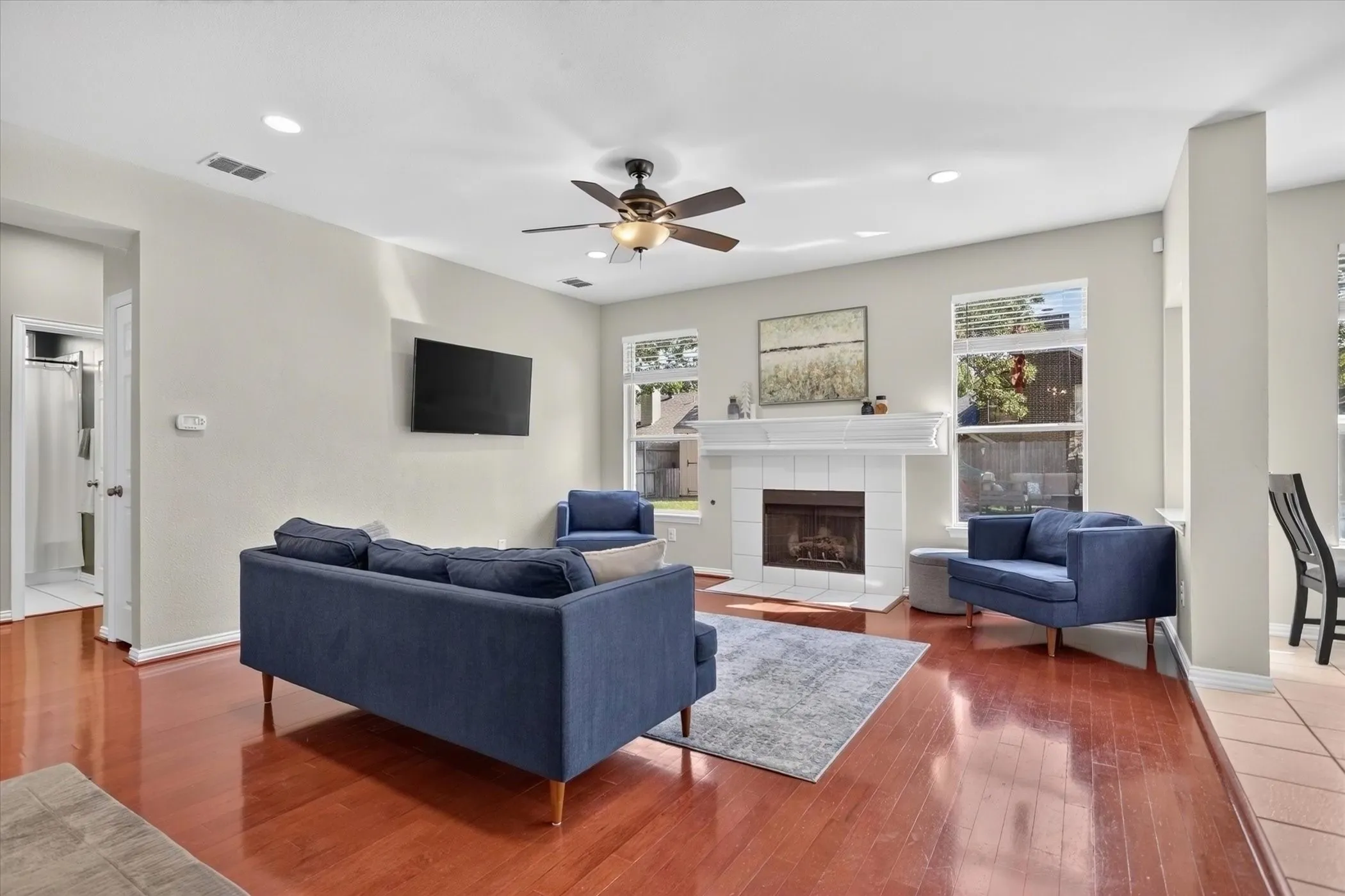 Living area featuring wood-finish flooring, a fireplace with a white tile surround and mantel, large windows, and a ceiling fan with integrated lighting