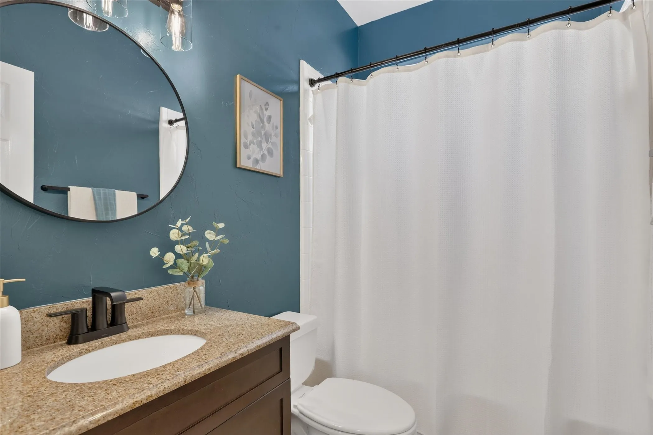 Vanity featuring a stone-composite countertop, undermount oval sink, and dark-finish cabinetry, complemented by a circular wall mirror and dark-finish hardware