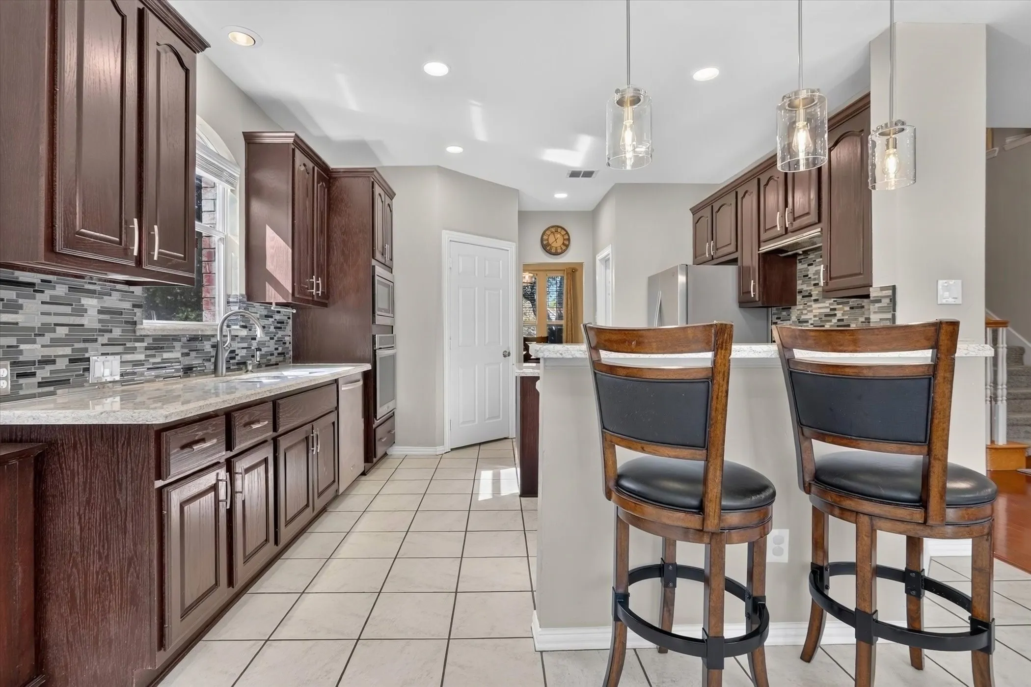 Kitchen featuring dark wood cabinetry, stone countertops, multi-tonal mosaic tile backsplash, stainless steel built-in appliances, and pendant lighting