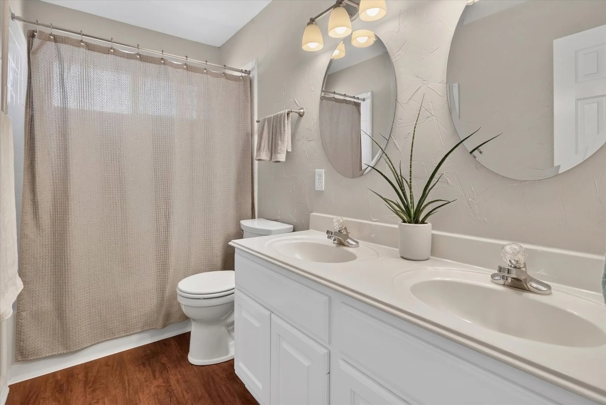 Bathroom featuring a dual-sink vanity with a solid-surface countertop, twin circular frameless mirrors, wood-finish flooring, and multi-bulb integrated lighting