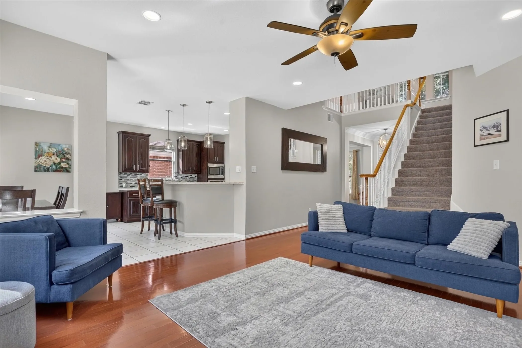 Open-concept interior featuring wood-finish flooring, recessed lighting, a ceiling fan, a kitchen island with pendant lighting, and a staircase with a wooden handrail