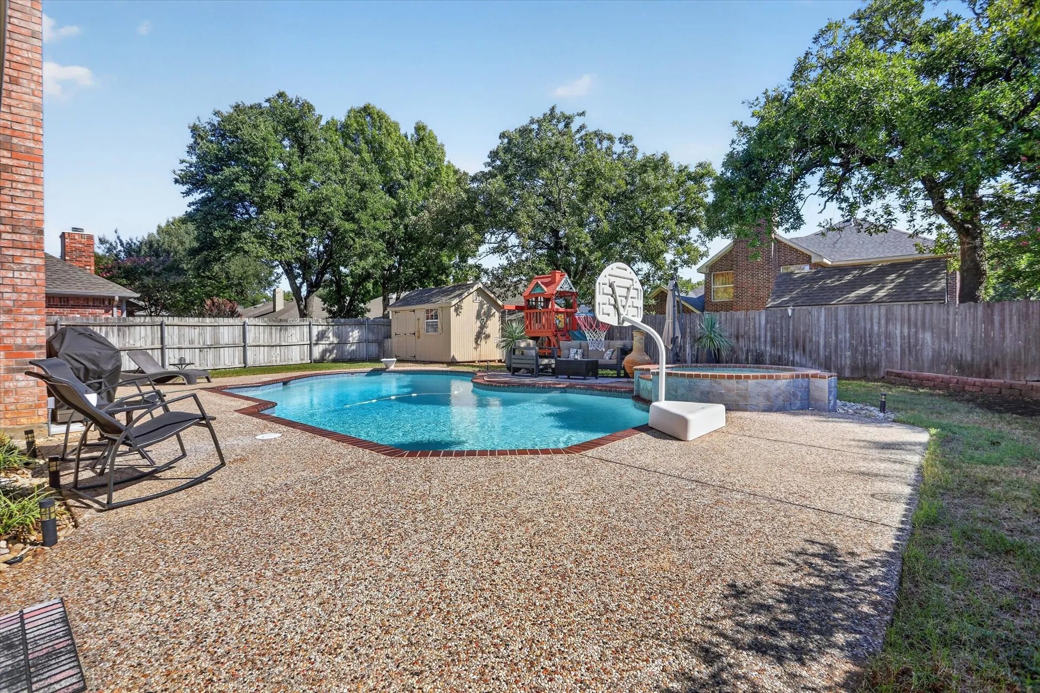 Backyard featuring an in-ground swimming pool with brick coping, an elevated stone-finished spa, aggregate concrete decking, and perimeter wood privacy fencing