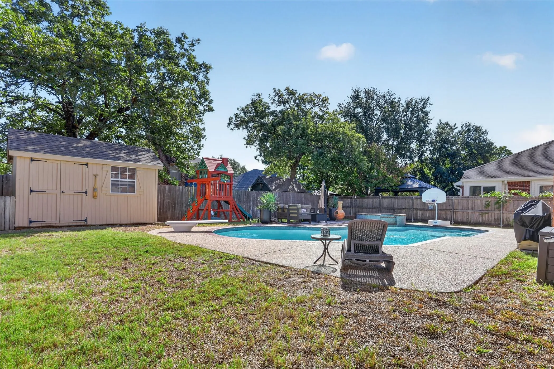 Backyard featuring an in-ground swimming pool with a diving board, aggregate patio surface, wood-paneled storage shed, and mature perimeter trees