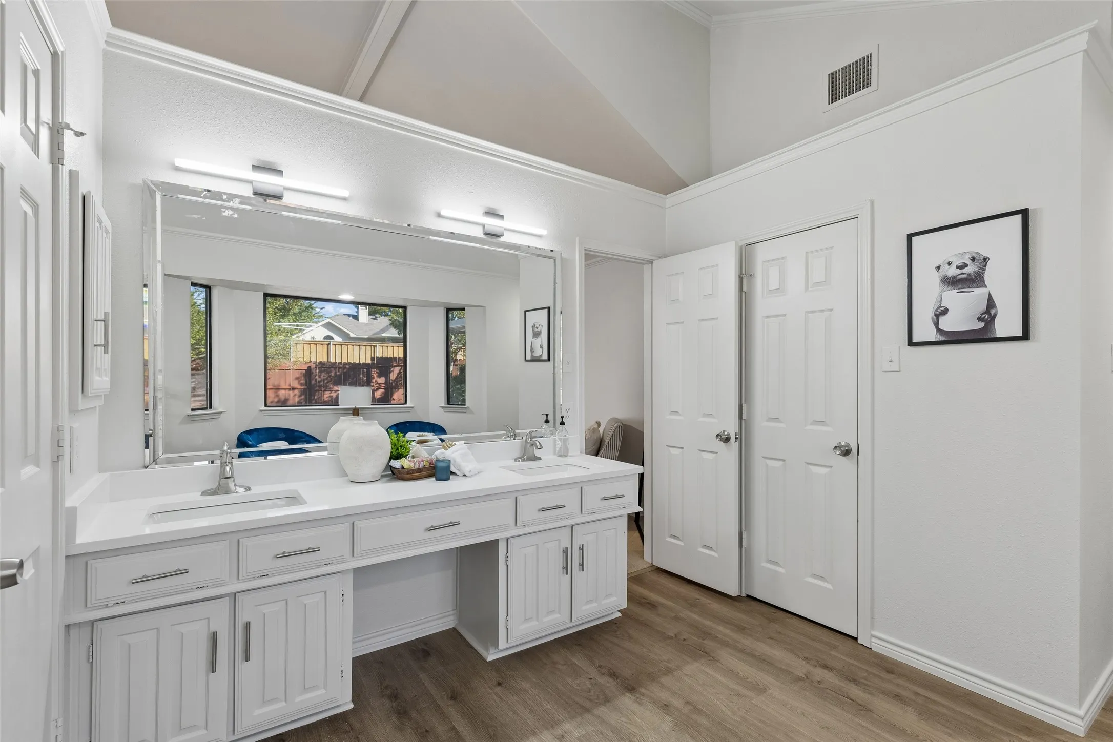 Bathroom featuring a dual-sink vanity with white cabinetry, light-toned countertops, a large full-width mirror, vaulted ceilings, and wood-finish flooring