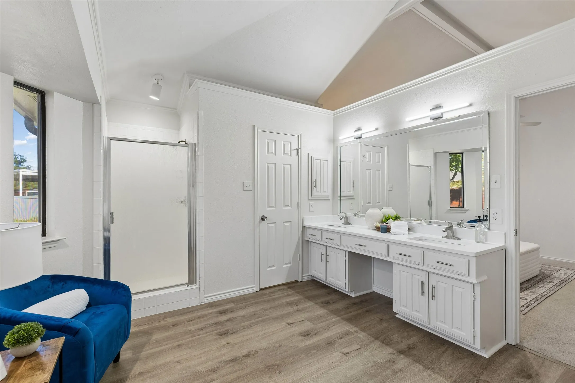 Vaulted ceiling featuring exposed beams, dual-sink vanity with white cabinetry, expansive wall-mounted mirror, walk-in shower with chrome enclosure, and wood-finish flooring