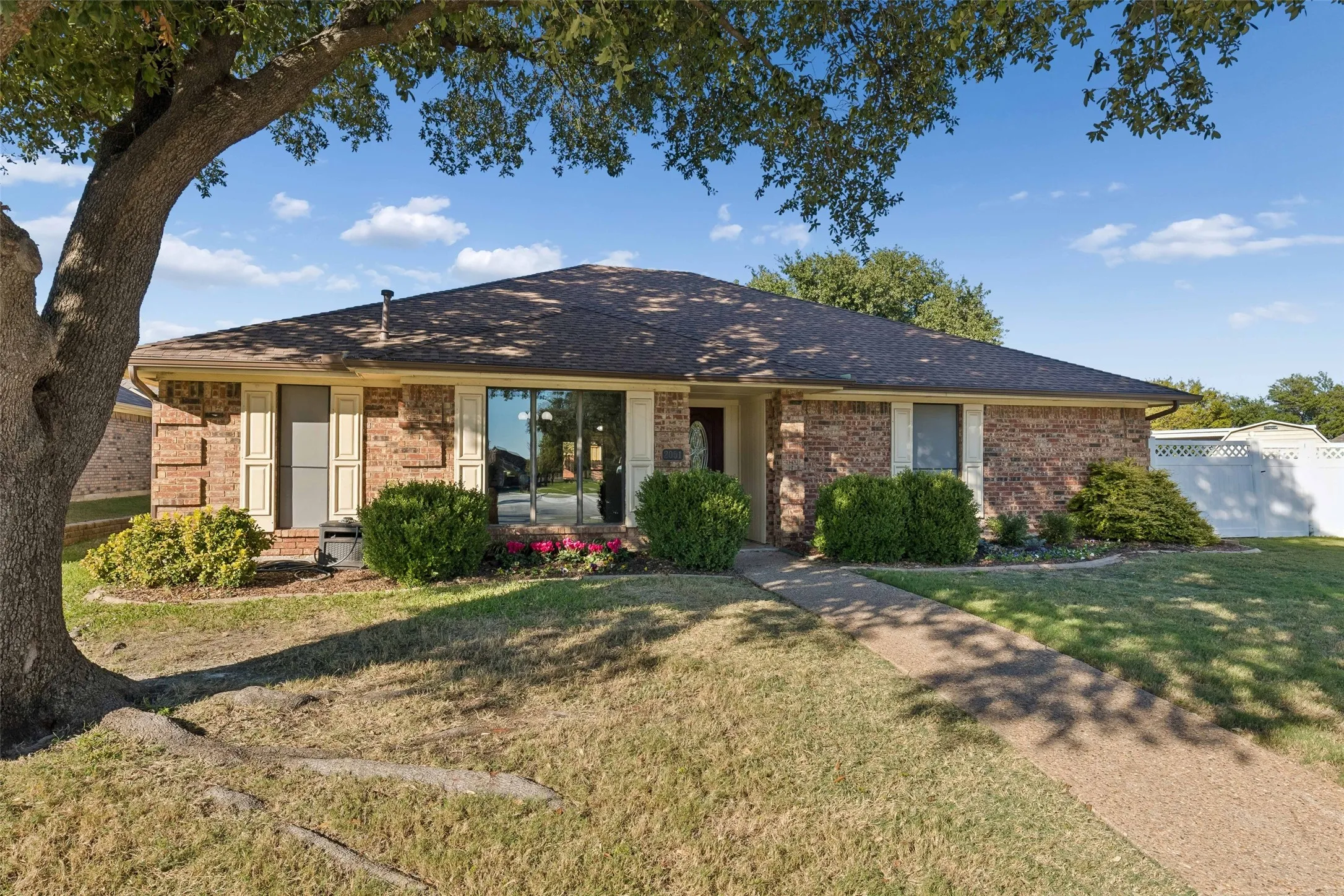 Single-story brick exterior featuring a hipped roof, contrasting window shutters, a concrete walkway, and established landscaping