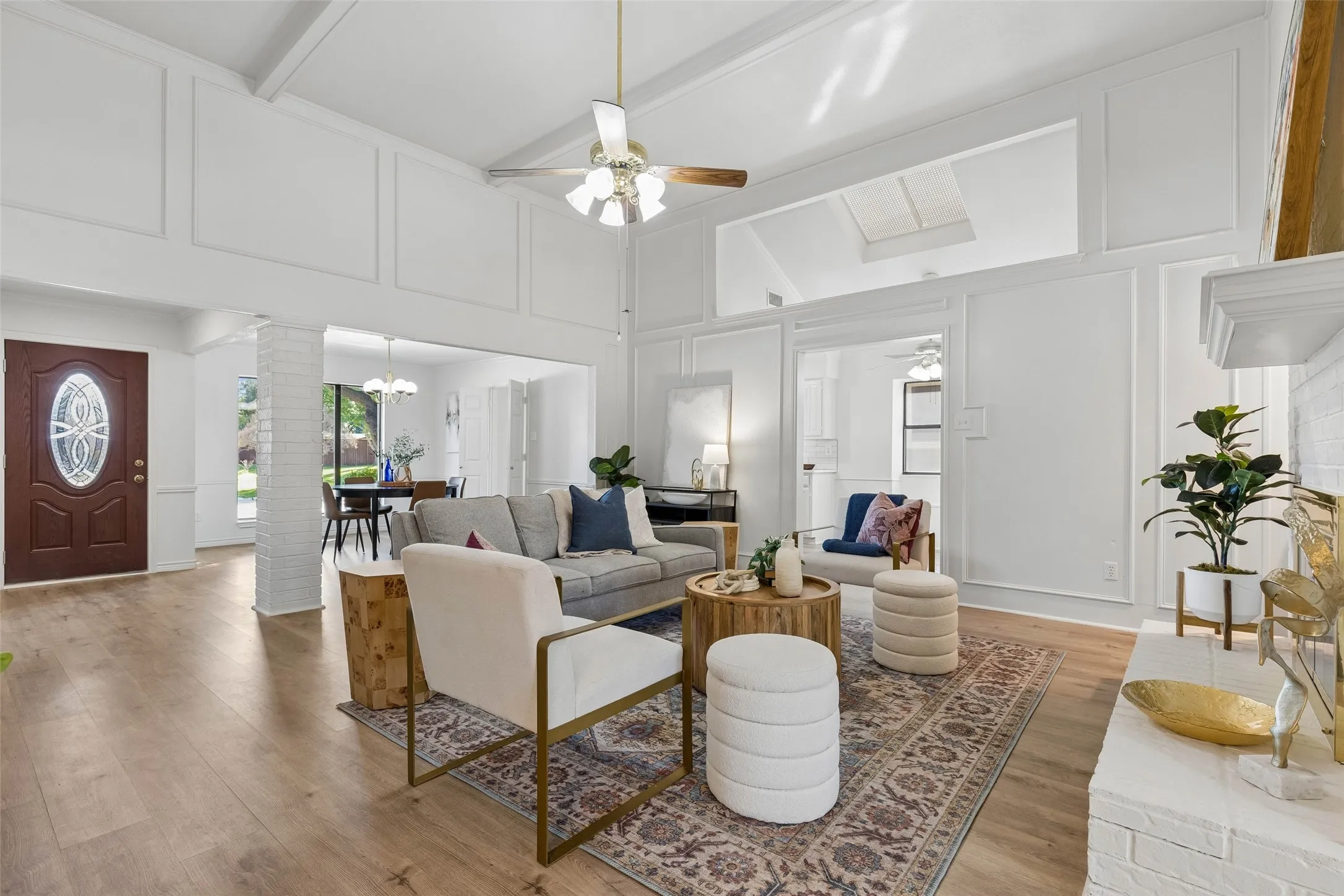 Vaulted ceilings featuring decorative wall paneling, wood-finish flooring, a white-painted brick fireplace, a skylight, and a ceiling fan